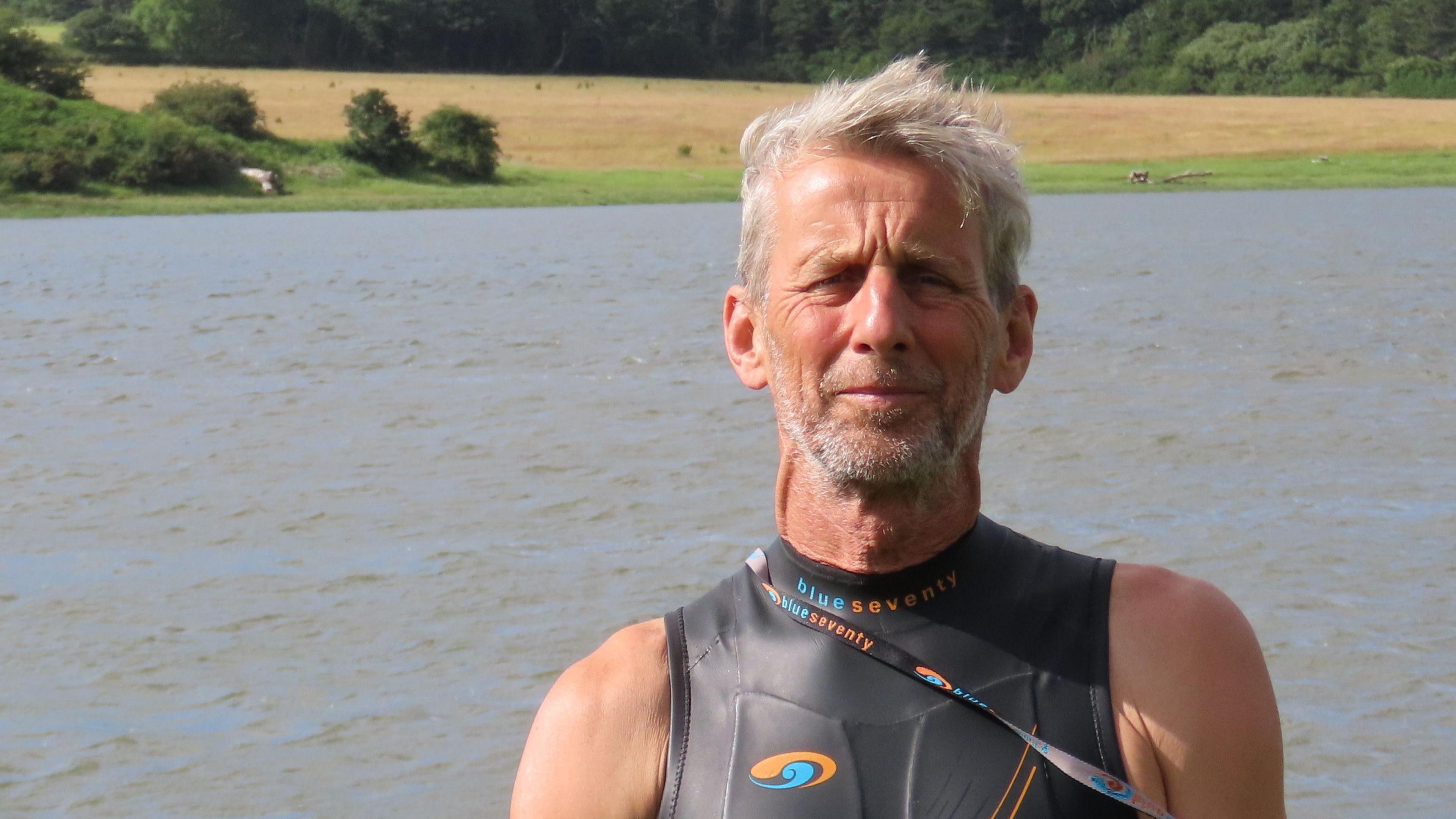 Ric Cooper of The Cleddau Project ready to go for a swim in the Western Cleddau river. He's standing in front of the river wearing a black, sleeveless wetsuit. He has grey short hair and grey stubble.
