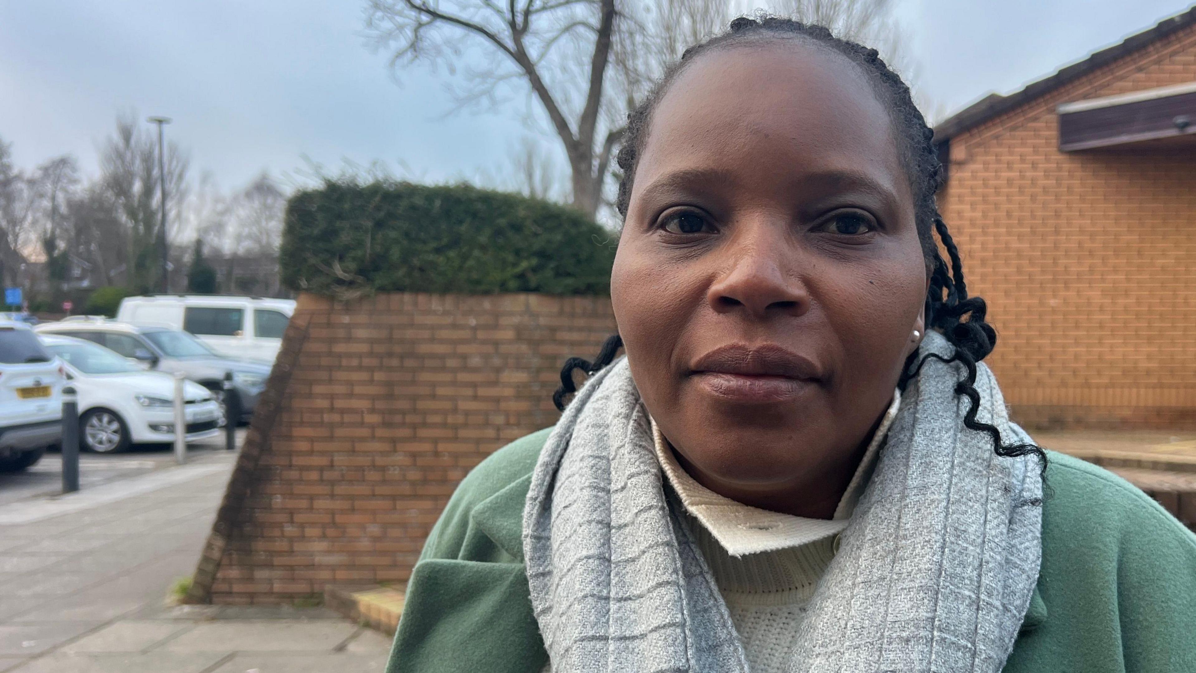 Martha Mhepo who has braided dark hair and is wearing a green coat and grey scarf. She is stood in front of a brick wall. next to a car park.