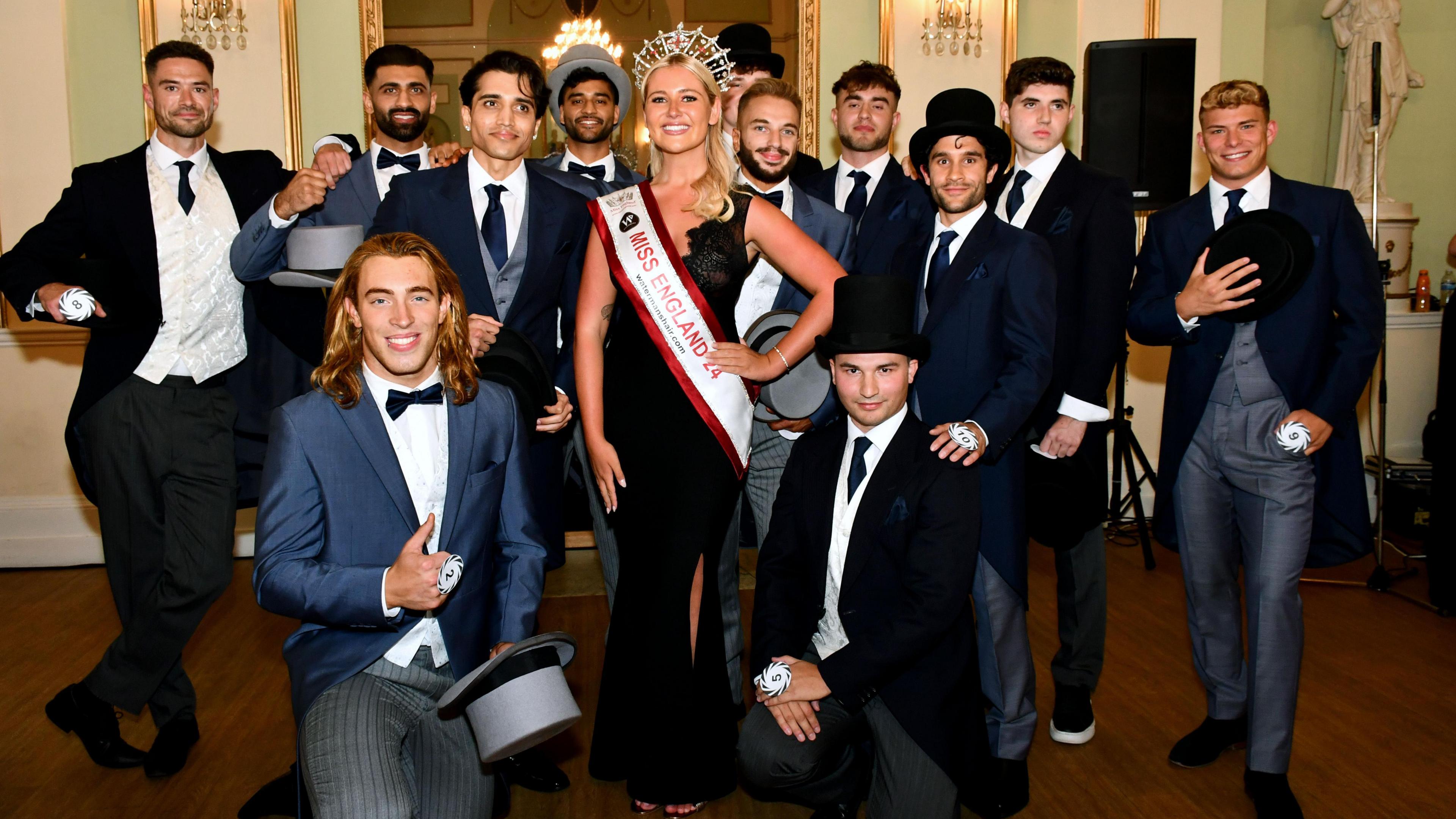 Bal poses for a picture with the other finalists, all in top hat and tails. Miss England is in the centre, wearing her crown, sash, and long black dress.