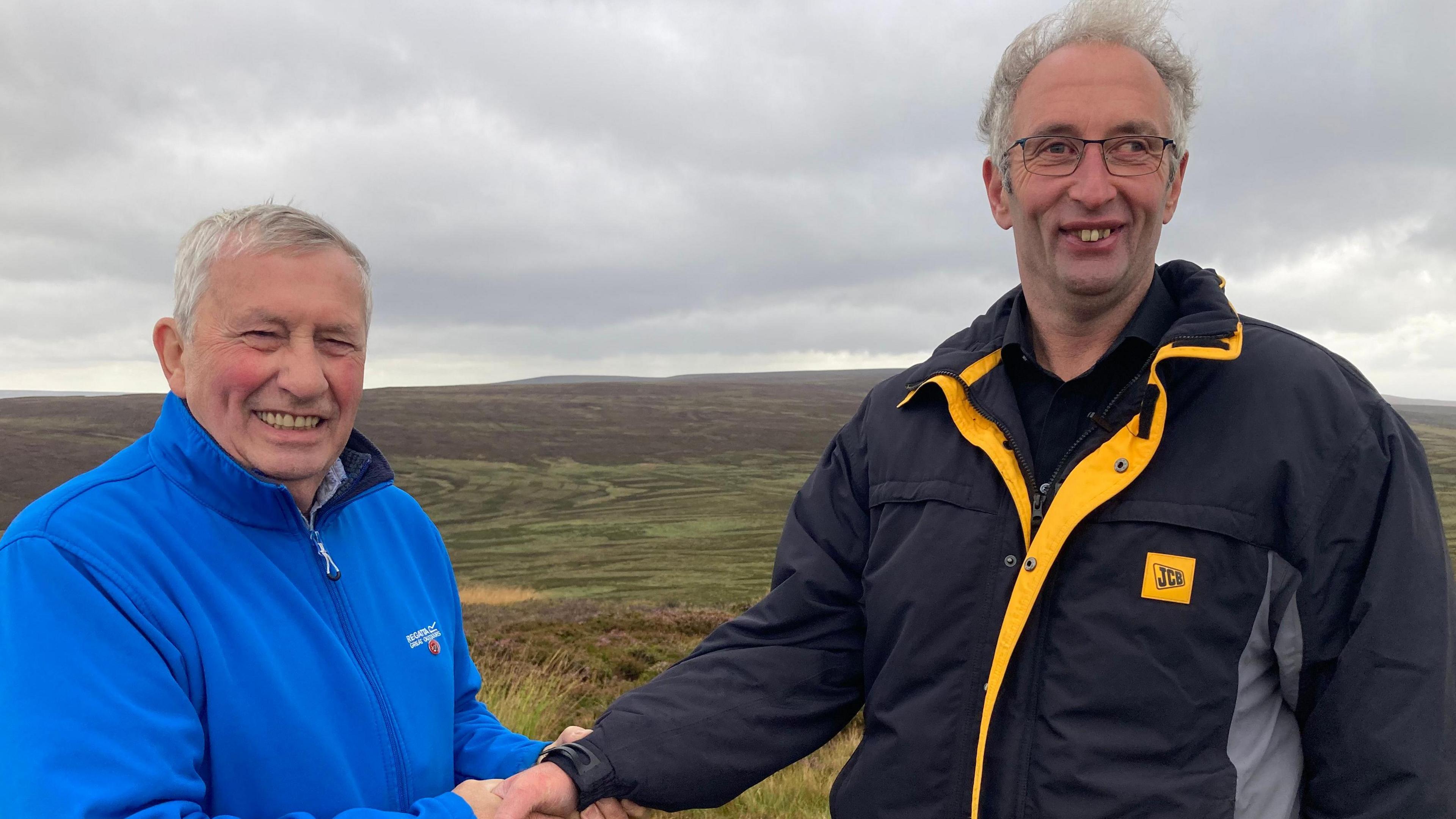 Earl Parker on the left in a blue fleece shaking hands with Ashley Hird in a black jacket with the Yorkshire Dales National Park in the background