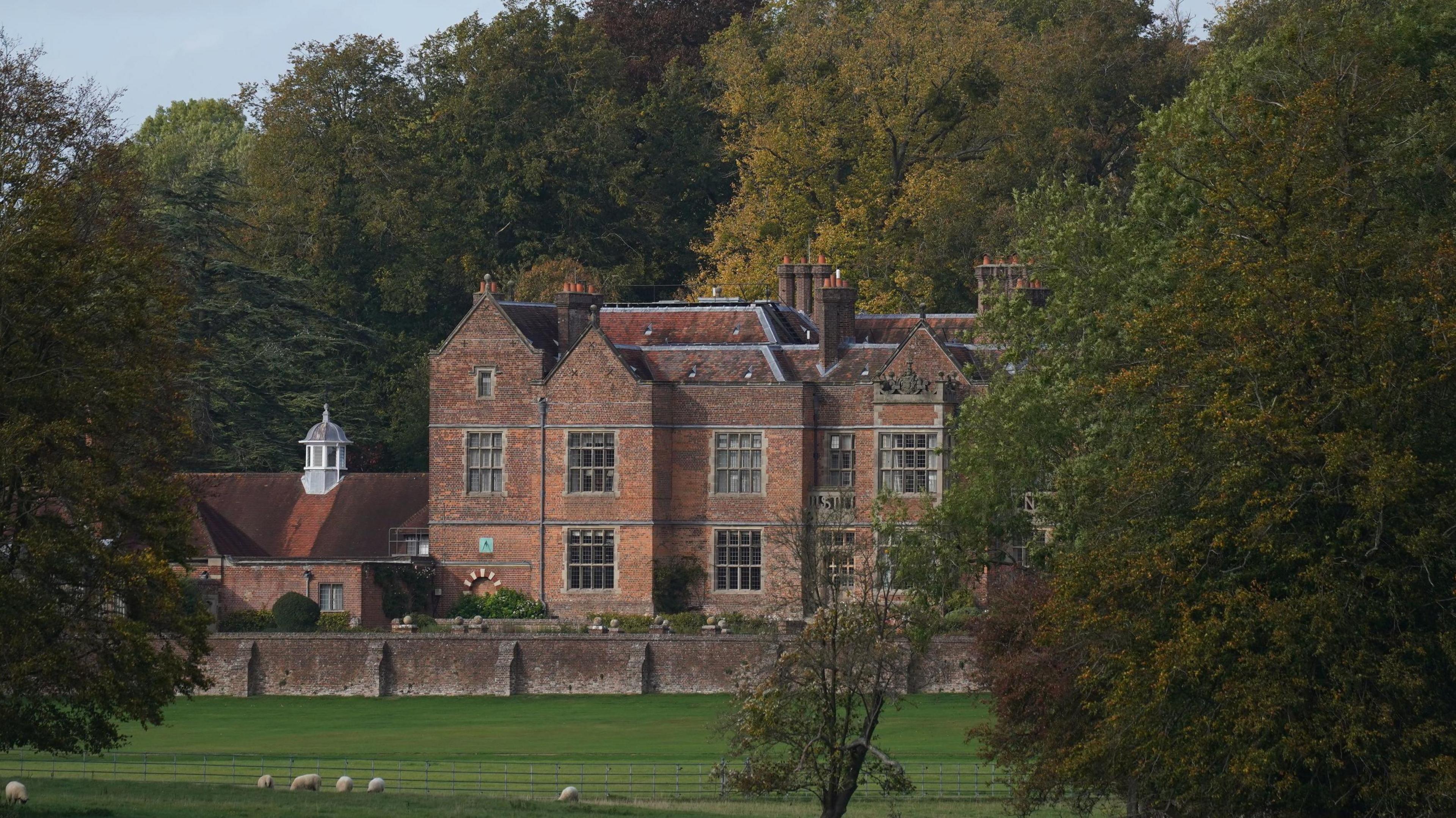 Chequers, in Buckinghamshire, a red bricked historical building, pictured from a distance with a long lens. The house is flanked by green trees and green lawns.