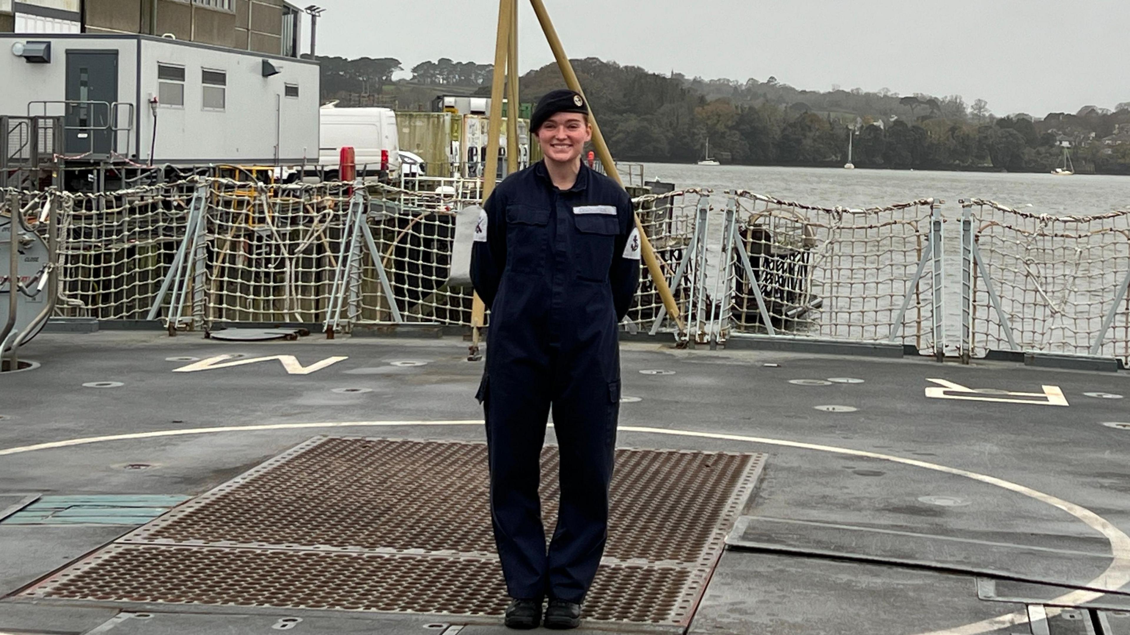 A woman smiling whilst standing onboard the HMS Richmond. The ocean and hills can be seen in the background. Grey skies.