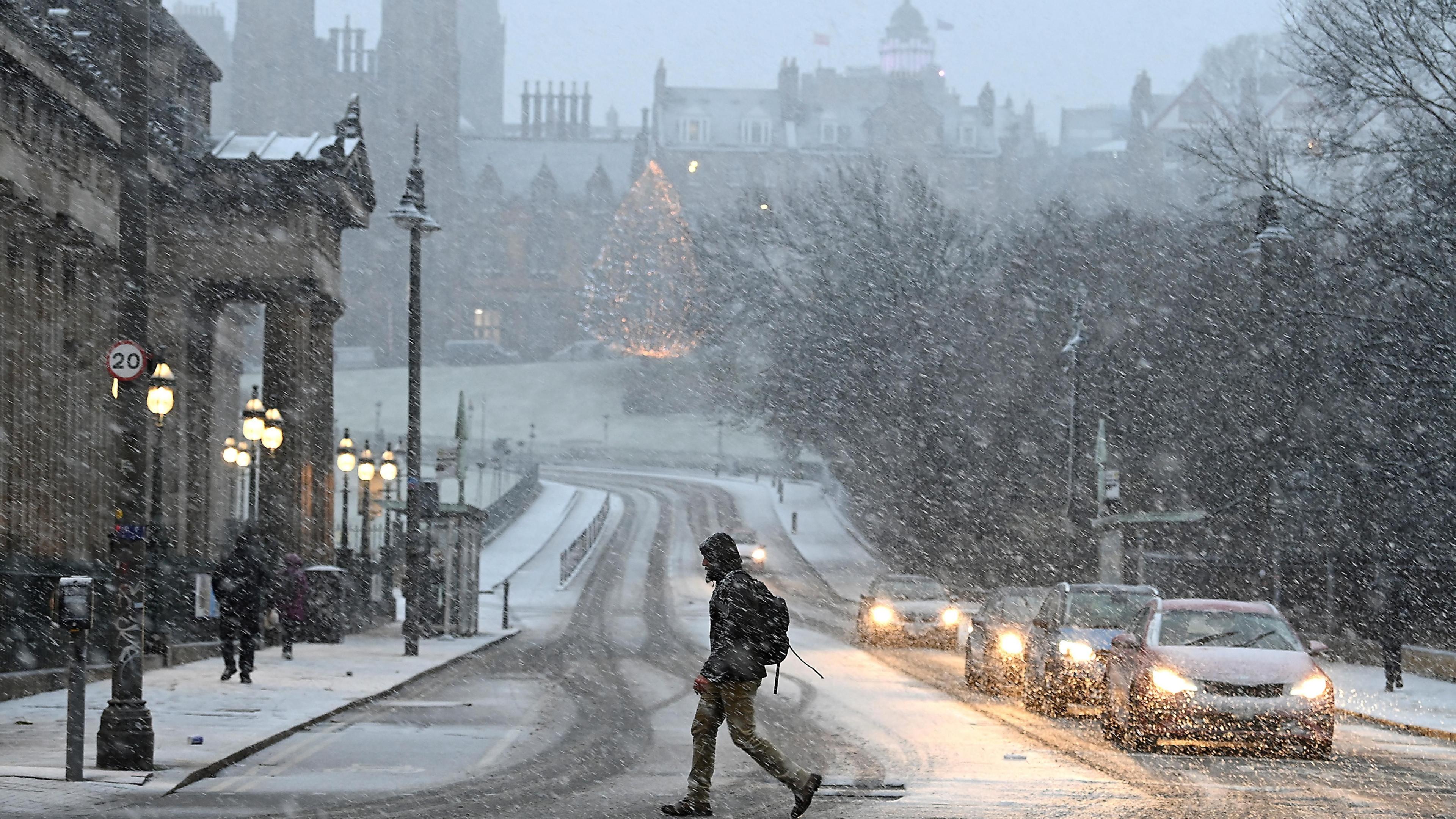 A man crosses a snowy junction with Princes Street in Edinburgh with Christmas tree in the distance during Storm Bert