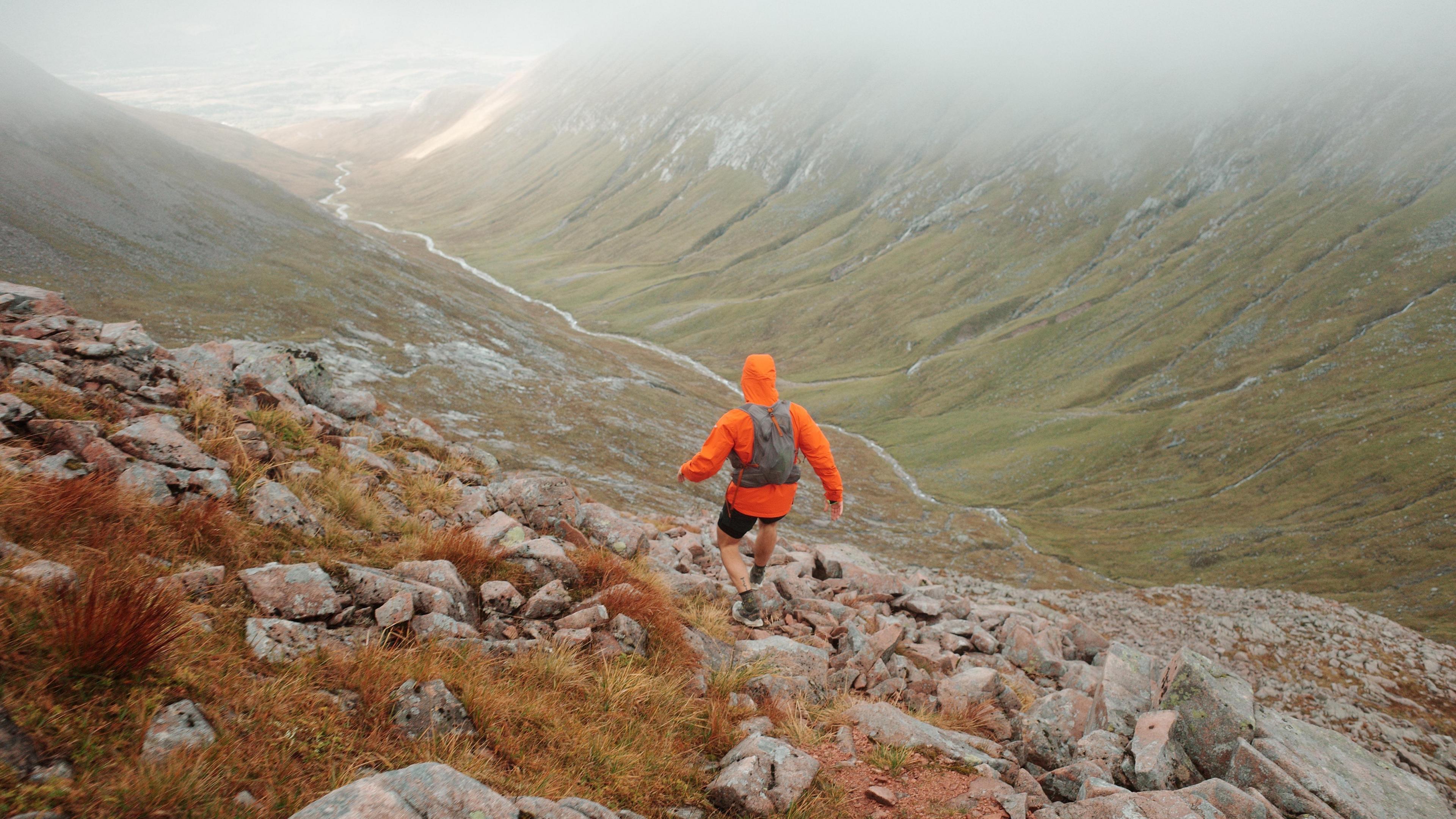 James Norbury walking through a mountainous area. He is pictured with his back to the camera, wearing a bright orange jacket, shorts, hiking boots and a backpack. The location is rocky. 