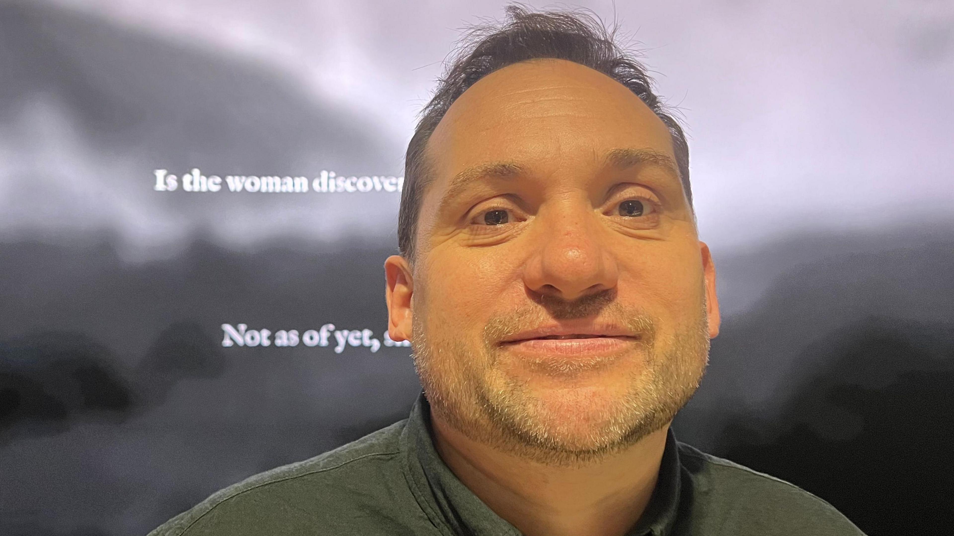 A man in a grey shirt with brown hair looks at the camera.