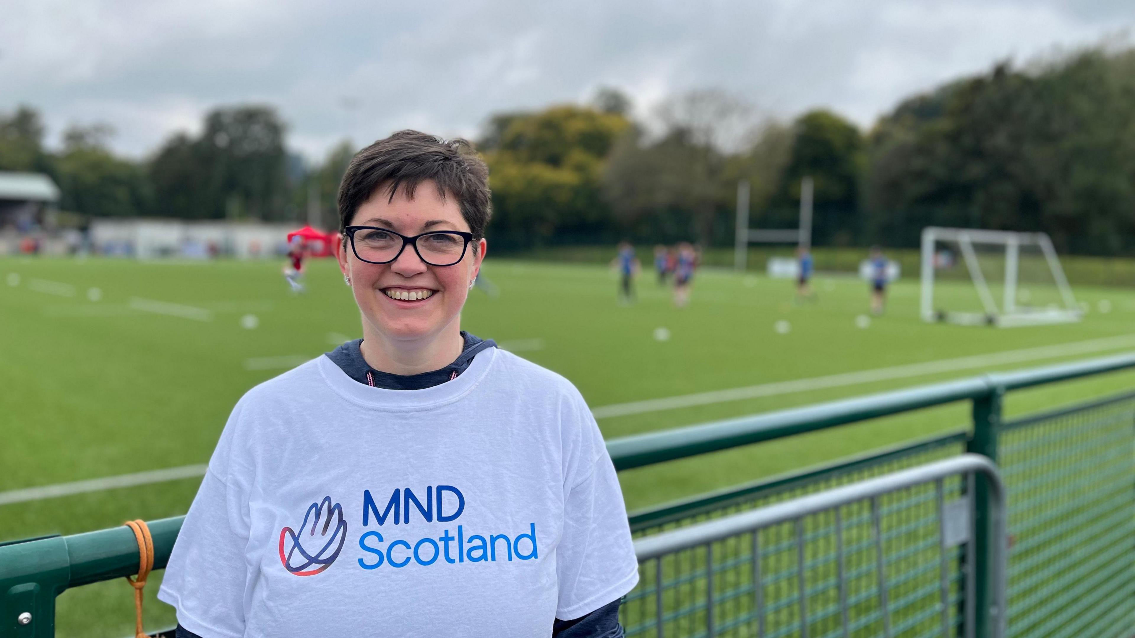 A woman in an MND Scotland T-Shirt standing in front of a football pitch