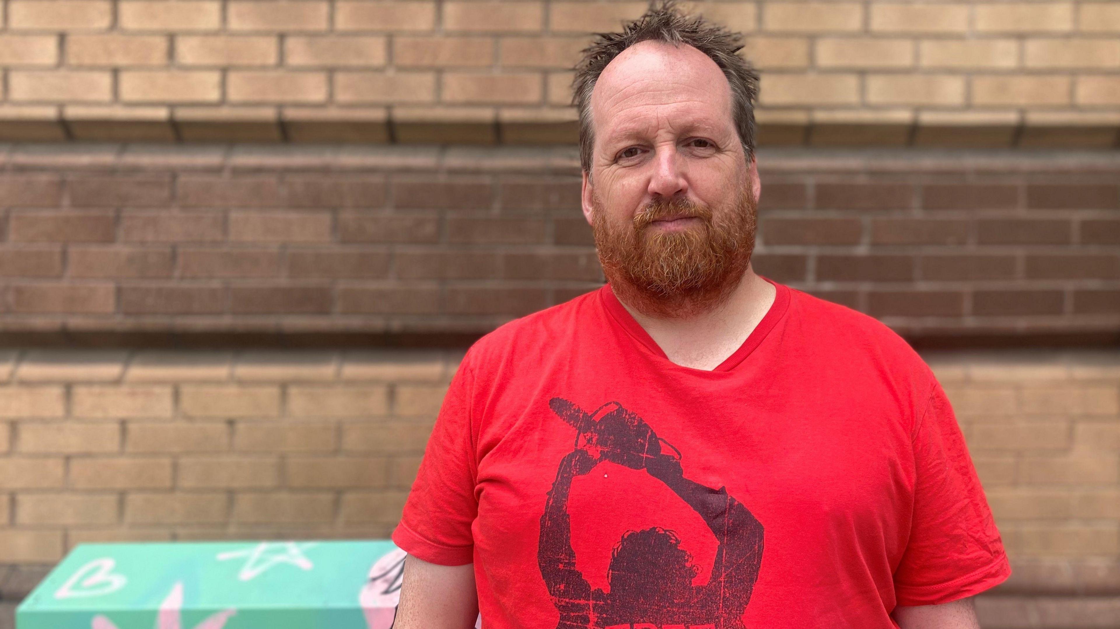 Adam is wearing a red t-shirt and is standing in front of a wall and a painted junction box which has been painted light green and the covered in light pink symbols like stars and hearts. Adam has light brown receding hair and ginger coloured beard. 