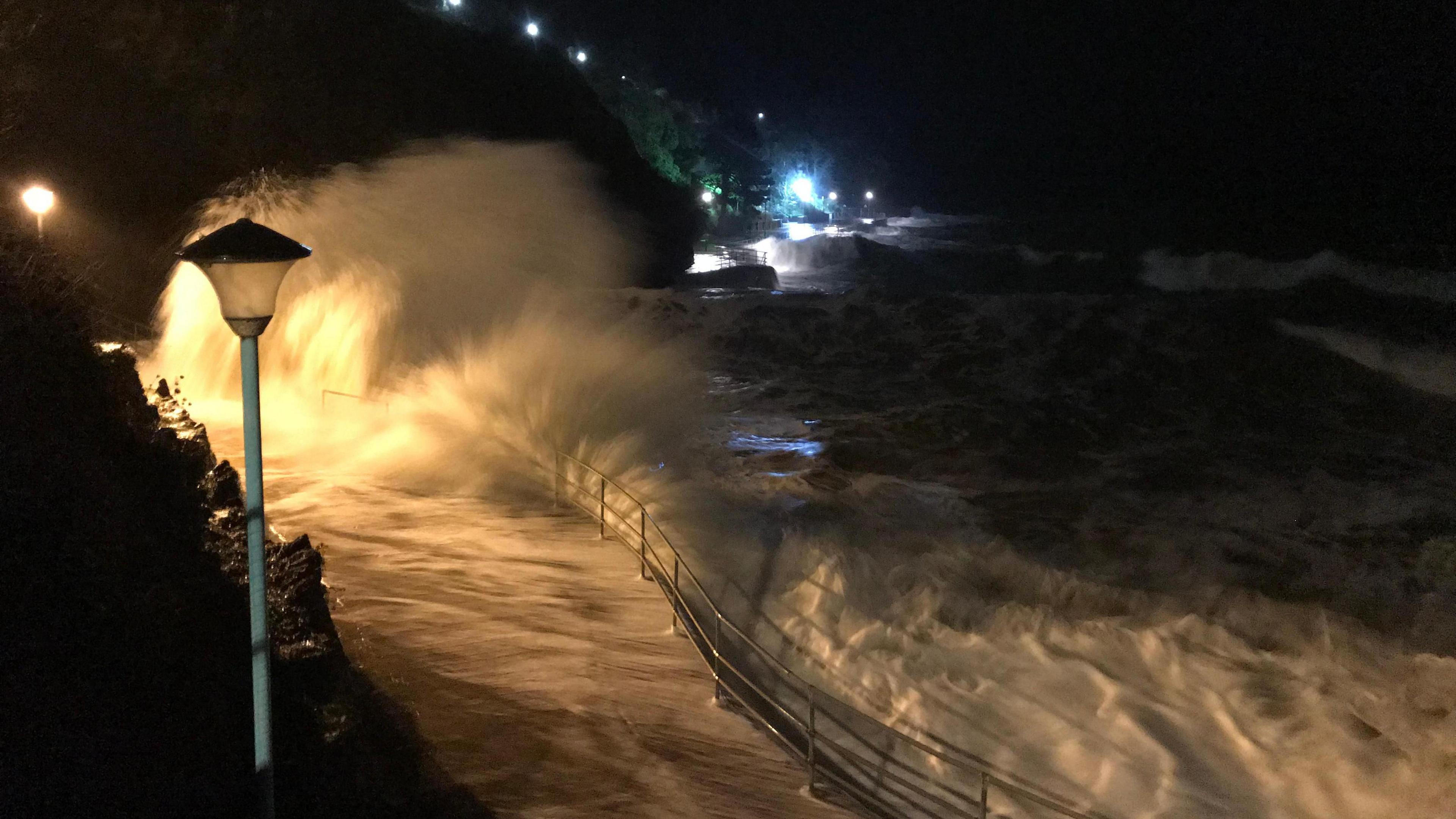 A waves crashes into the coastline in Goodrington at night. A walkway with railings on the side by the sea is covered in water. A streetlight, which is switched off, is in the foreground.