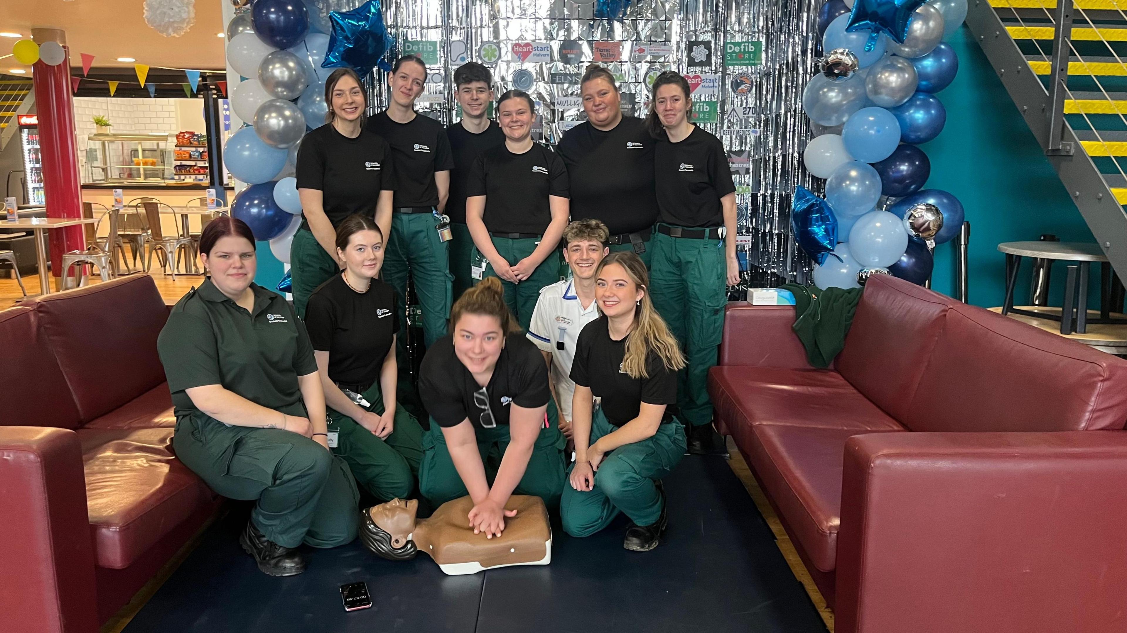 A group picture of paramedic students dressed in a black T-shirt and green trousers. The are all smiling positioned outside a blue balloon arch