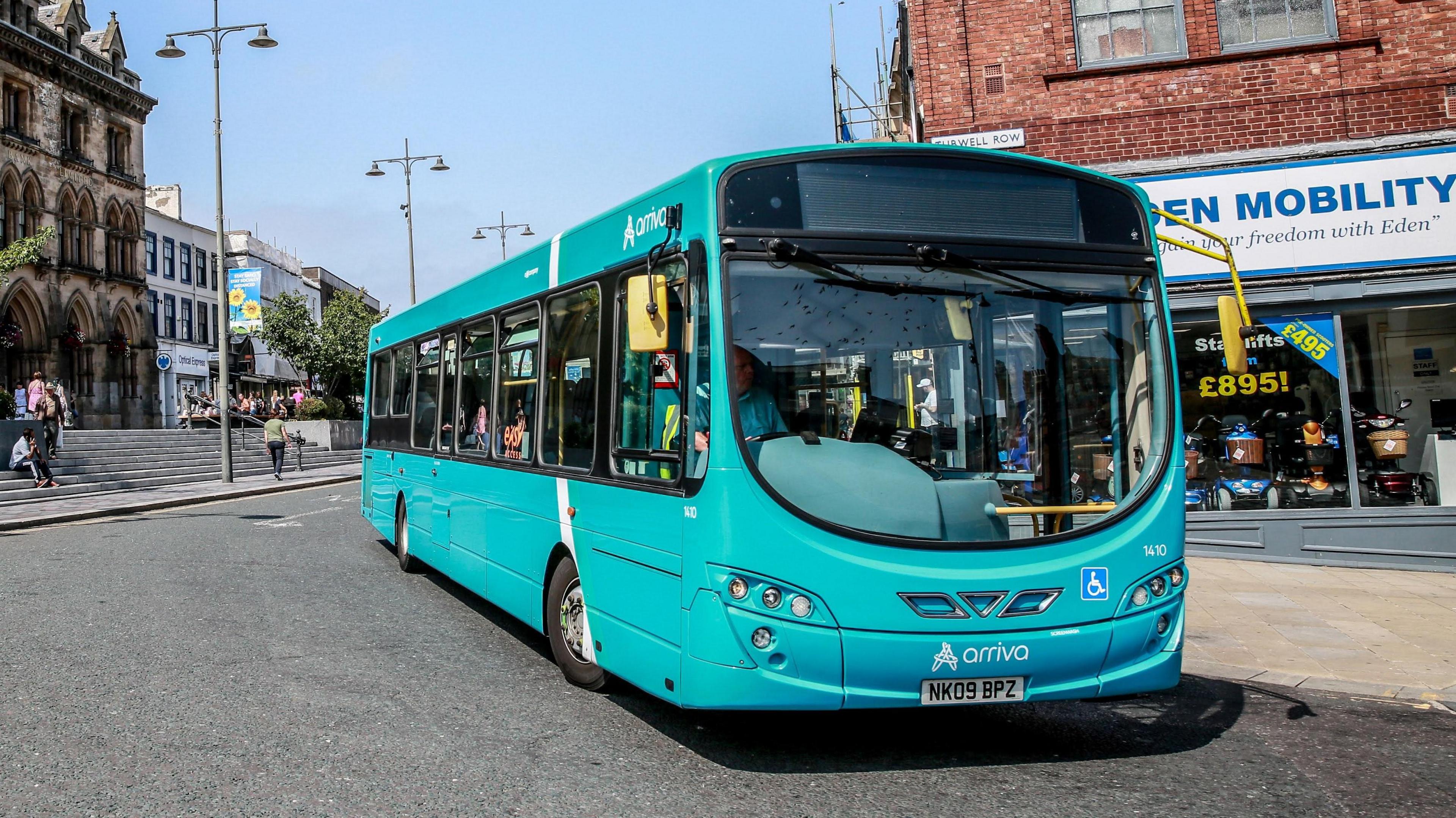A blue Arriva bus waits on a wide corner outside a mobility shop. There is no destination banner up but a driver is sat inside the bus.