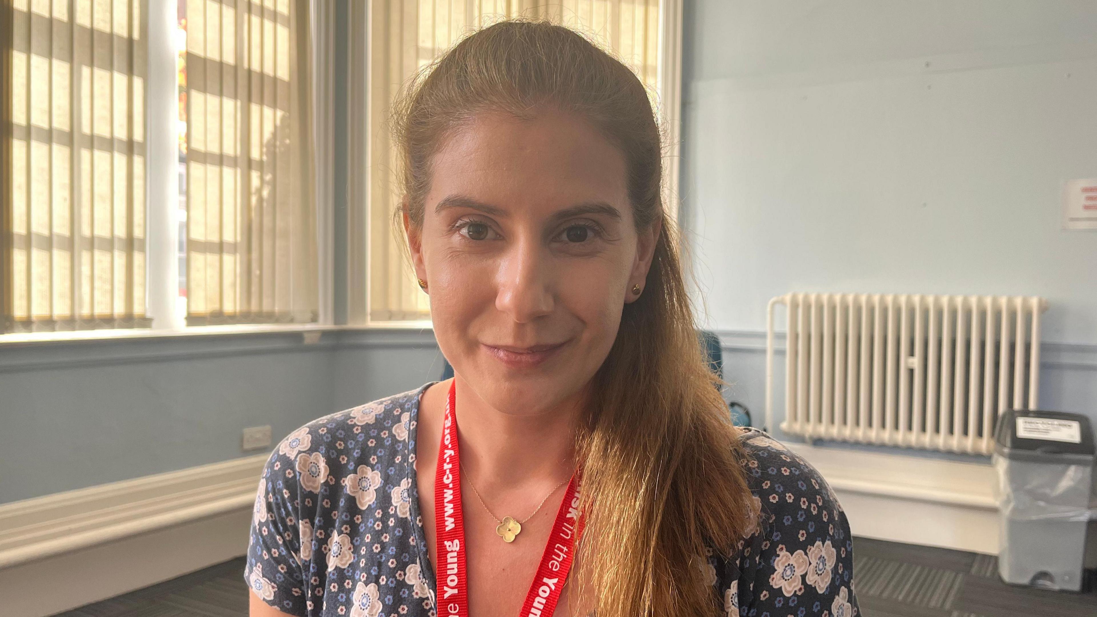 A head and shoulders shot of a woman smiling at the camera. She has long brown hair tied back in a ponytail and is wearing a floral dress and a red lanyard. She is sat in a room painted a light blue with large windows and blinds.