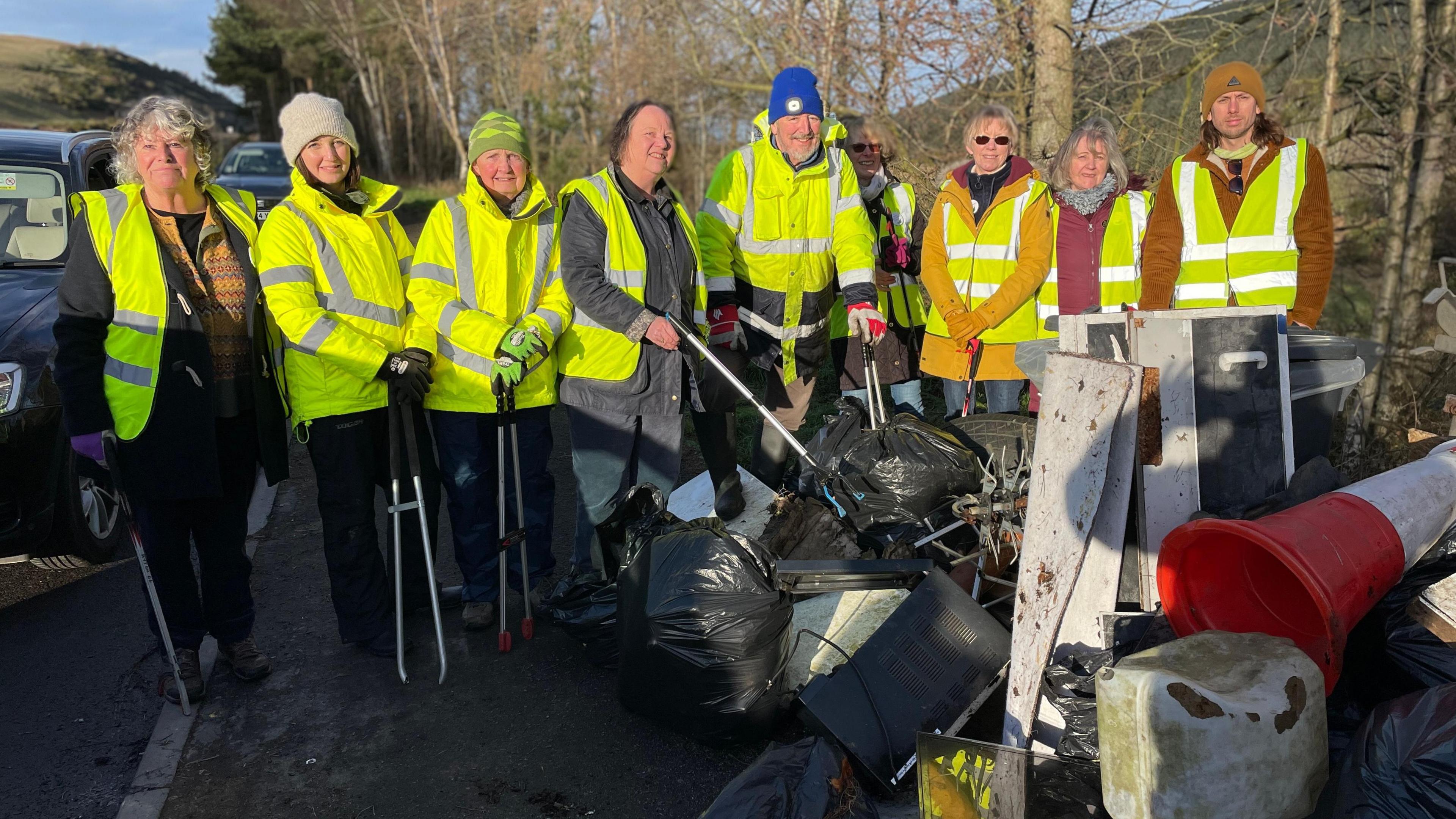 A total of nine people in high visibility jackets stand behind a pile of black bags and other discarded items.