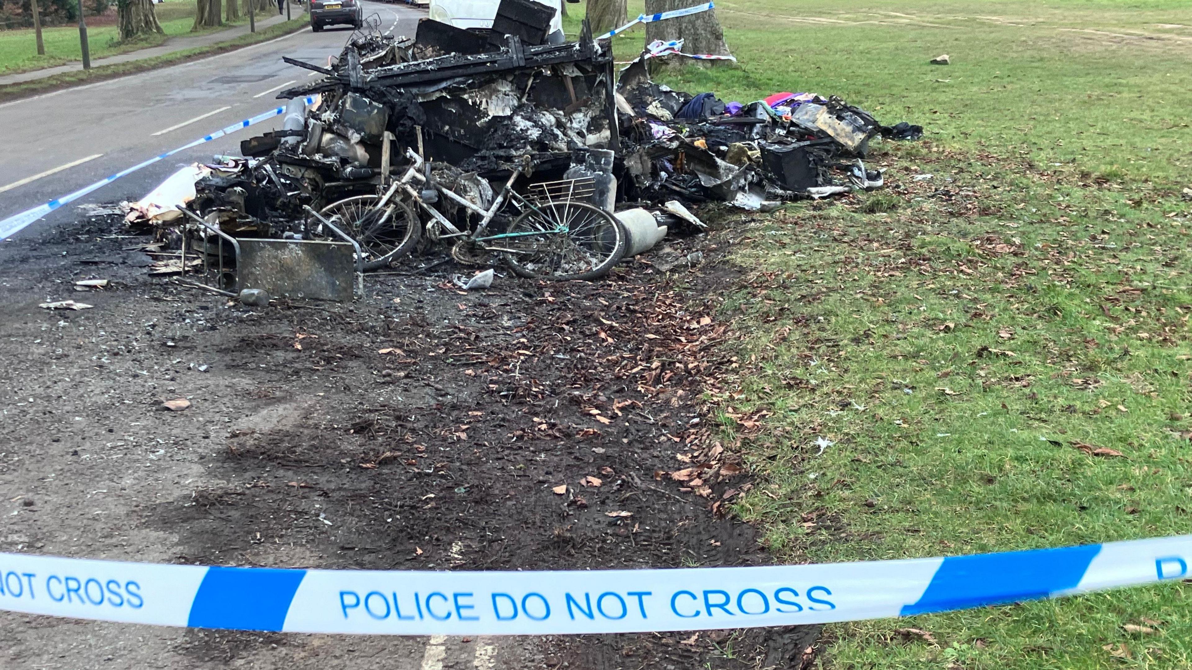 Police tape in front of a pile of burnt items which were destroyed in the fire next to a patch of grass.