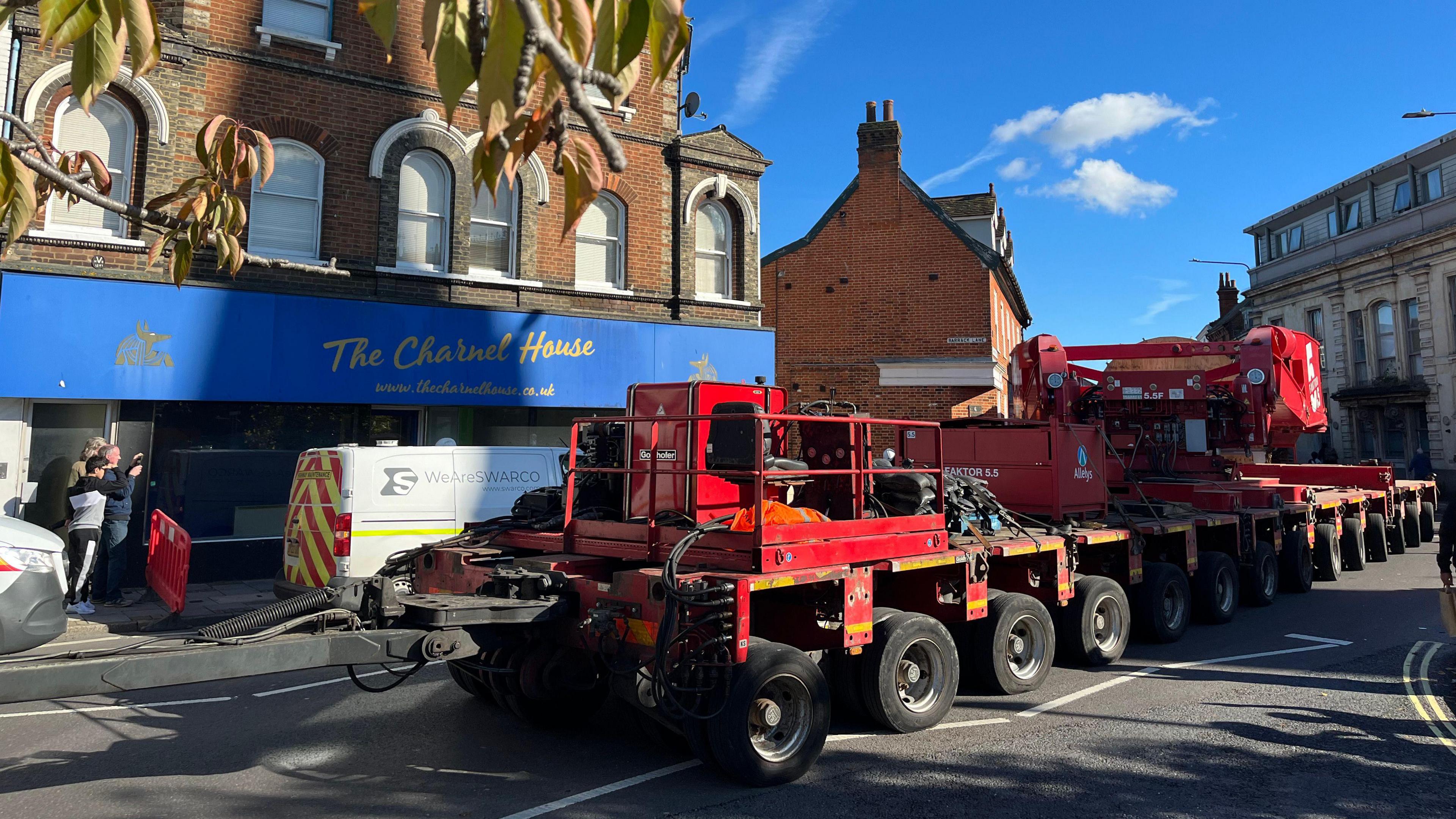 An abnormal load of a large red truck with more than 12 wheels, traveeling down a high street with shops in the back 