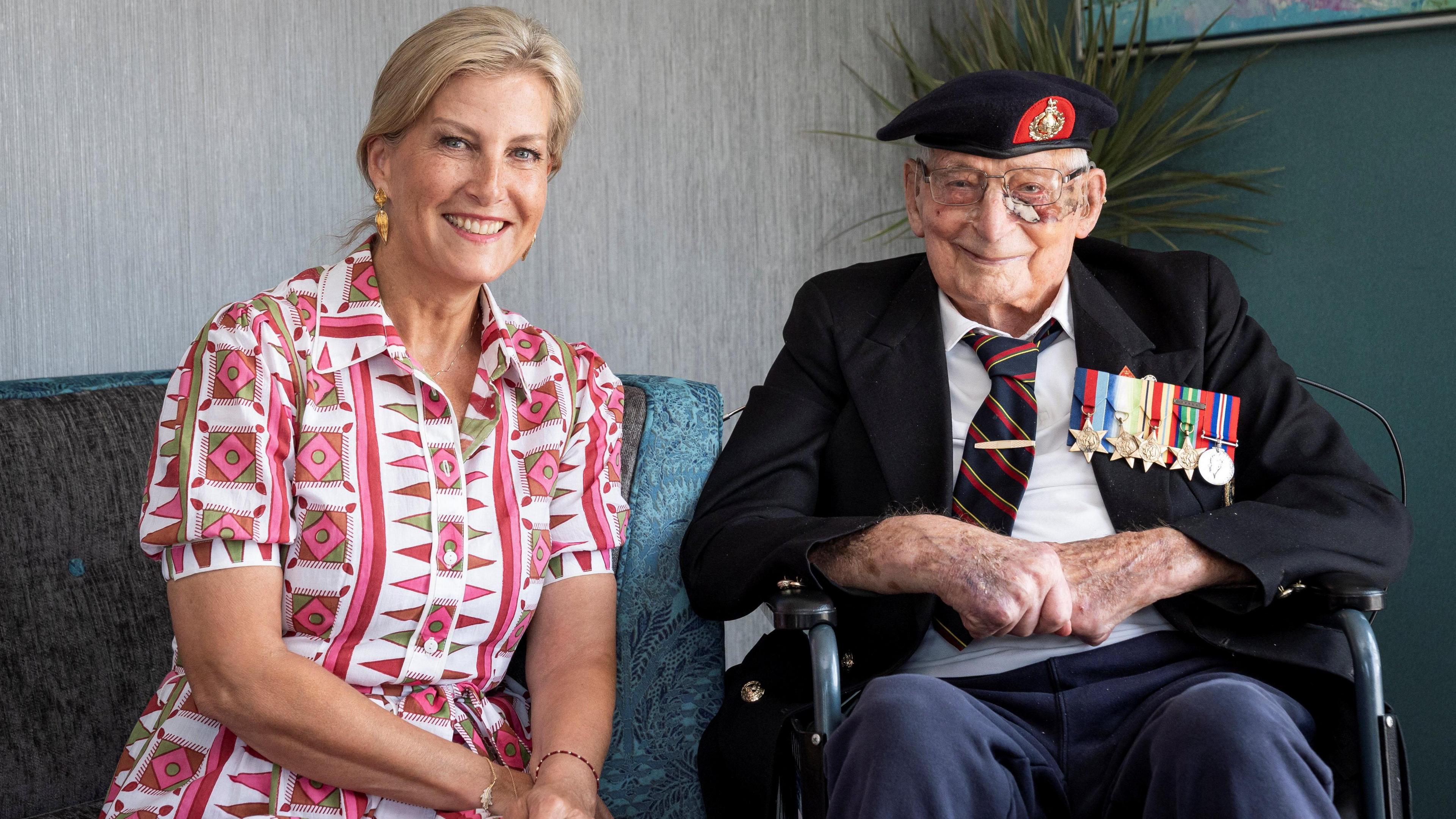 The Duchess of Edinburgh sat next to James "Jim" Wren. She is blonde and is wearing a white and pink patterned dress. She is looking ahead and smiling. Mr Wren is an elderly man dressed in a shirt, tie, blazer and a Royal Navy hat. He has several medals on his jacket. He is sat in a wheelchair, looking ahead and smiling.