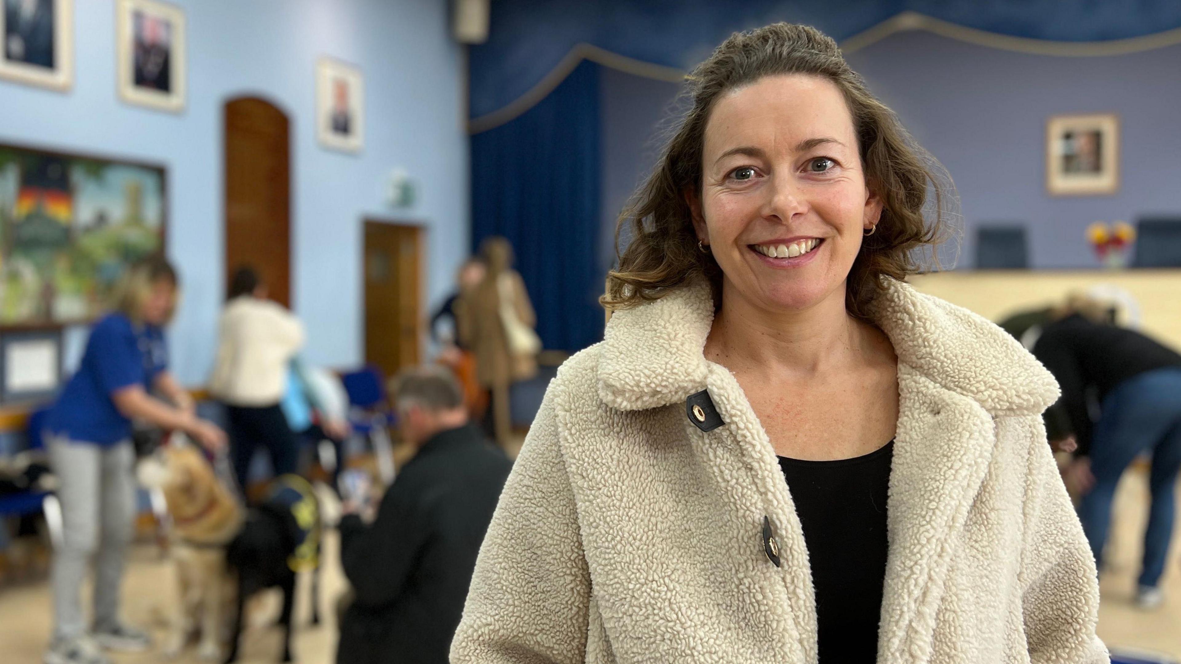 Elle is wearing a light-colored fleece coat stands in a hall with blue walls and framed pictures. In the background, several people interact with dogs near blue chairs and tables, with a stage visible at the far end.