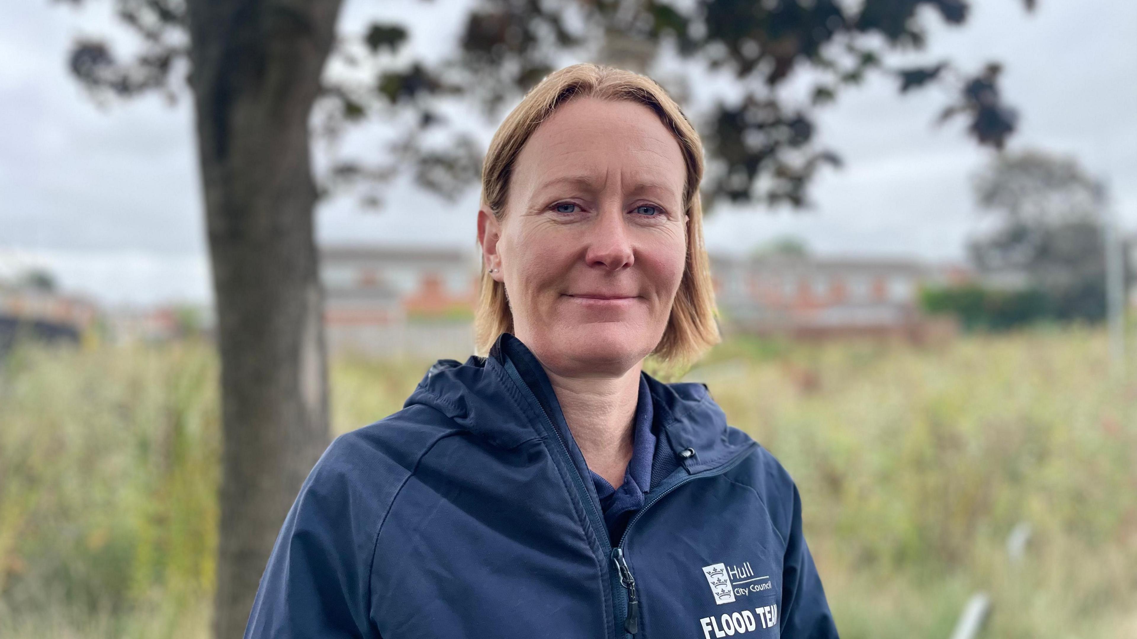 A woman with blonde collar-length hair parted in the middle smiles at the camera. She is wearing a blue waterproof jacket with a Hull City Council logo above the words Flood Team in white capital letters. The blurred background shows a tree and wild grass area in front of a row of modern houses.