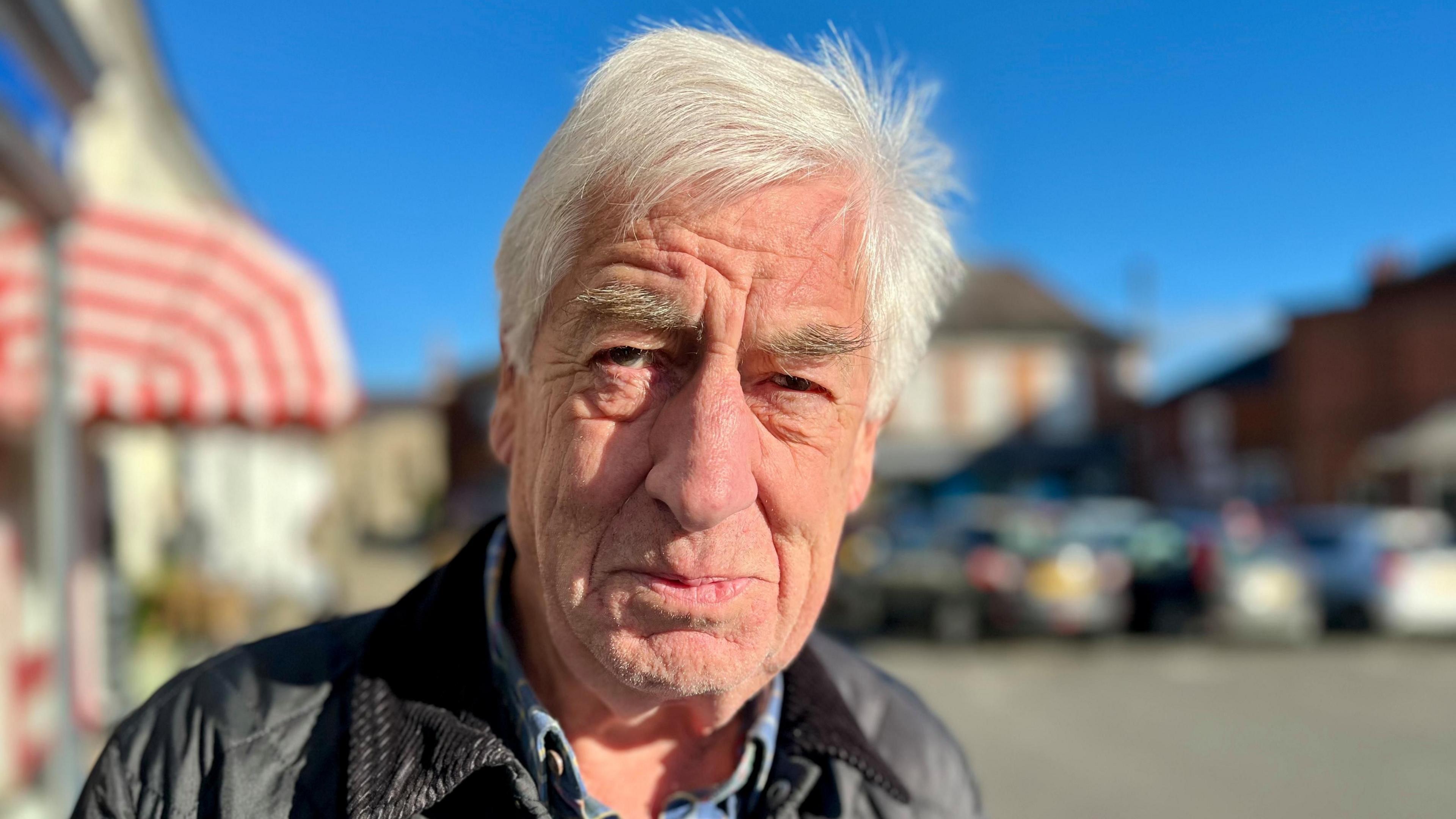 Keith Reeves has short white hair and is wearing a black leather jacket with a blue shirt underneath.
He is standing in Aylsham market place, smiling at the camera with shops and parked cars behind him.