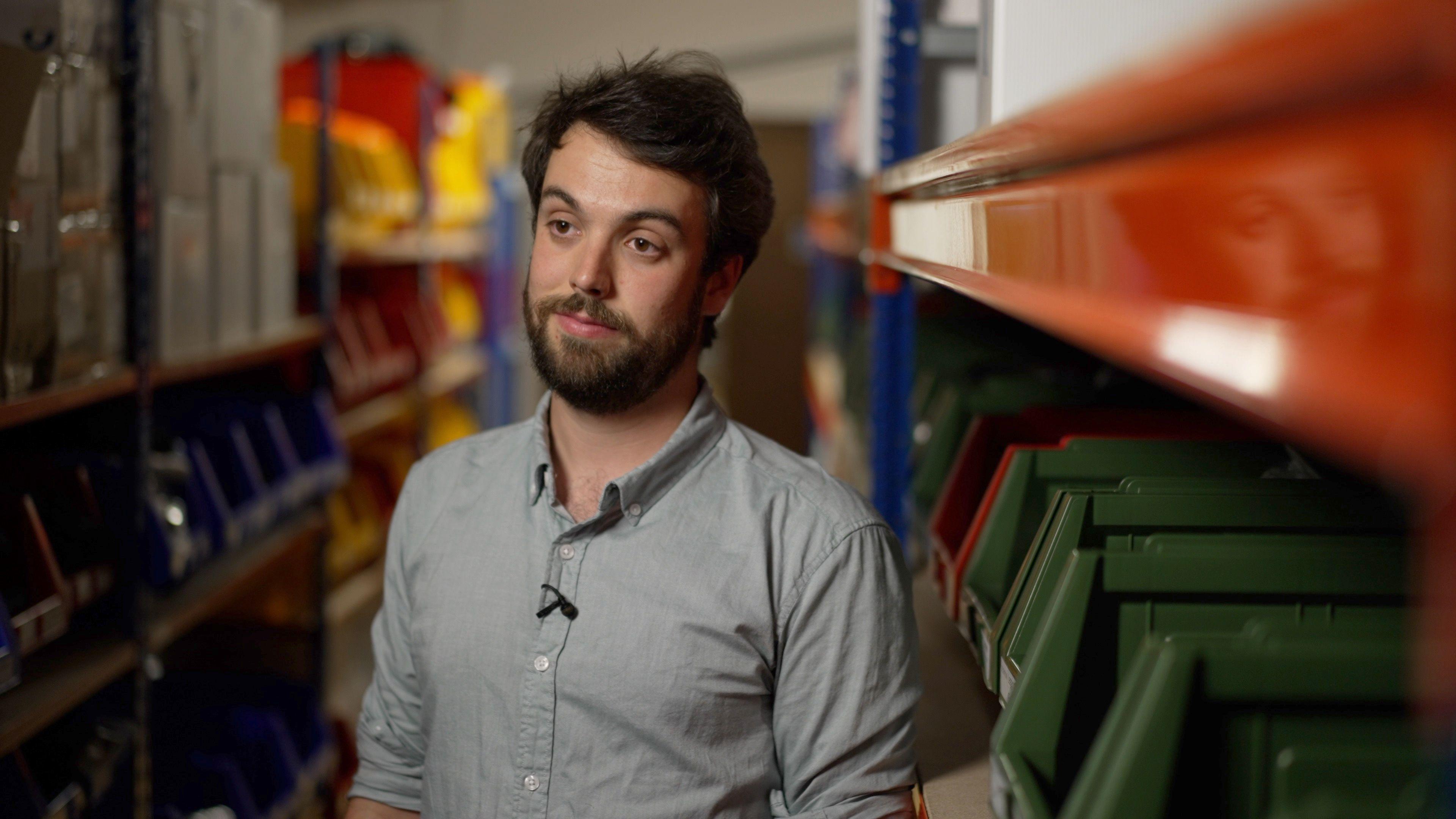 A man wearing a grey shirt stands in a corridor, with orange and green files around him.