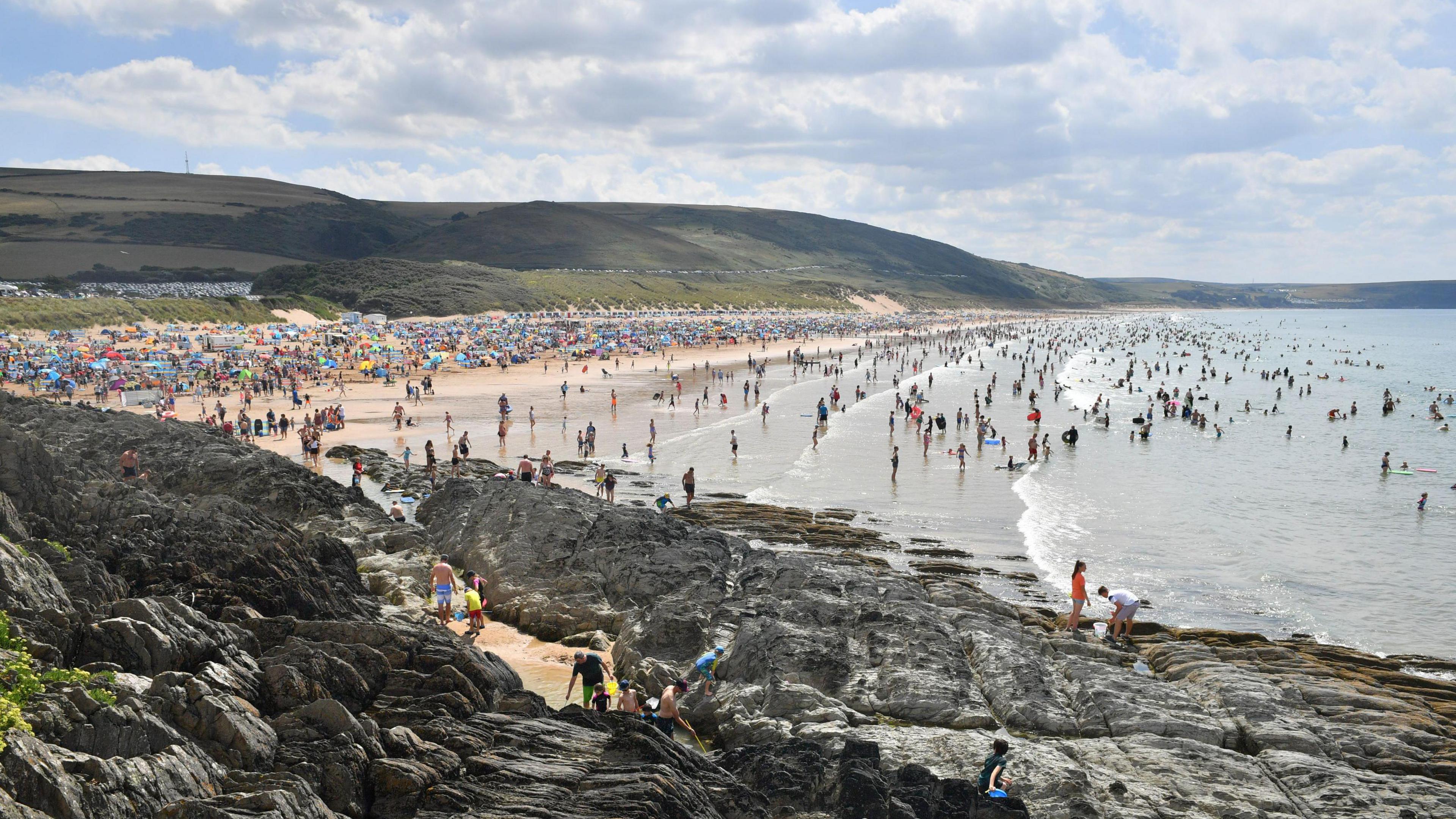 Woolacombe beach on a sunny day is packed with people and the sea is full of people wading and swimming. The grassy headland stretches down the coast and there are cars lined along a road on it. In the foreground there are rocks which a few people are standing on.