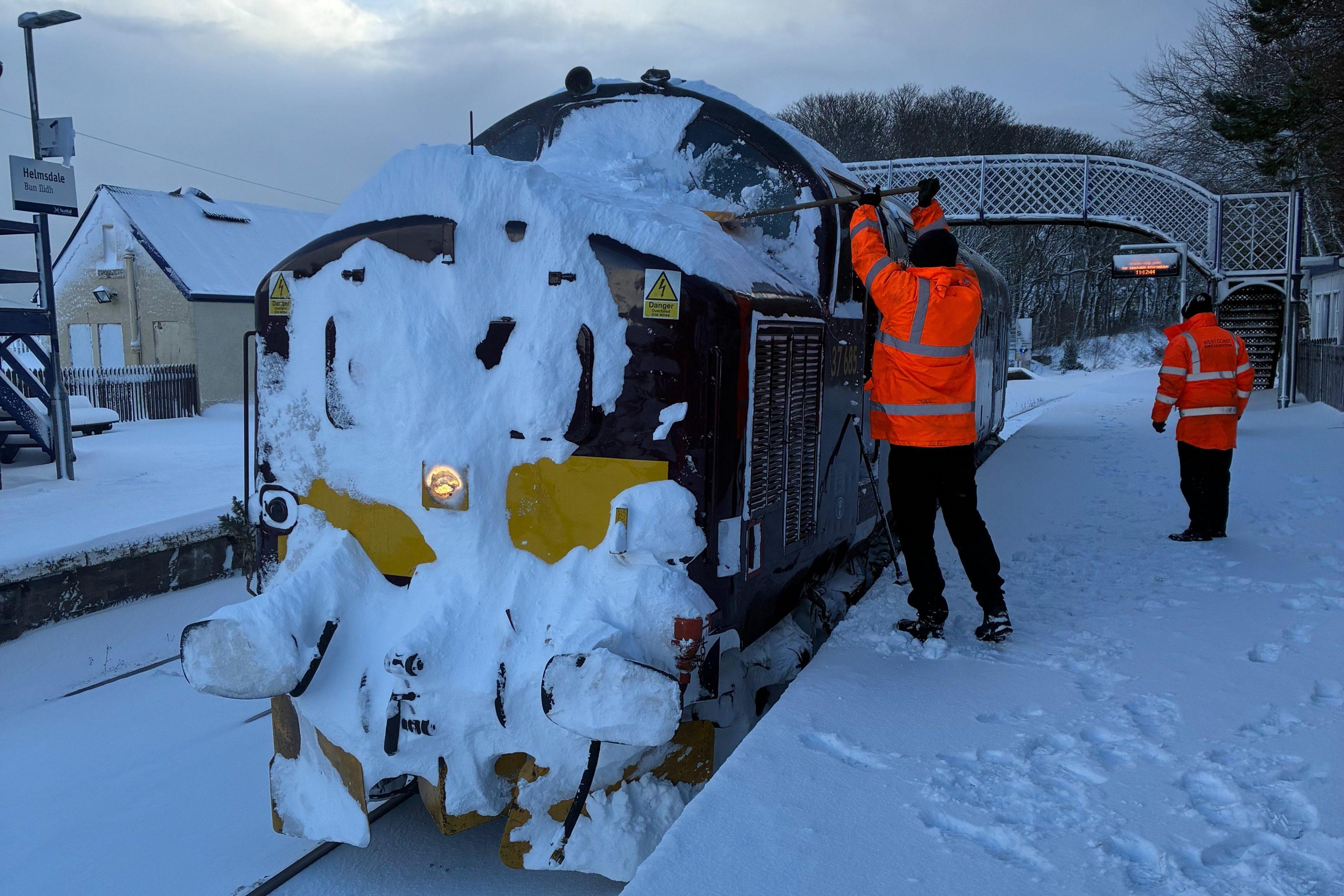Një tren me borë të mbuluar në pjesën e përparme në një stacion treni me dëborë. Platforma dhe shinat hekurudhore janë të mbuluara me borë.