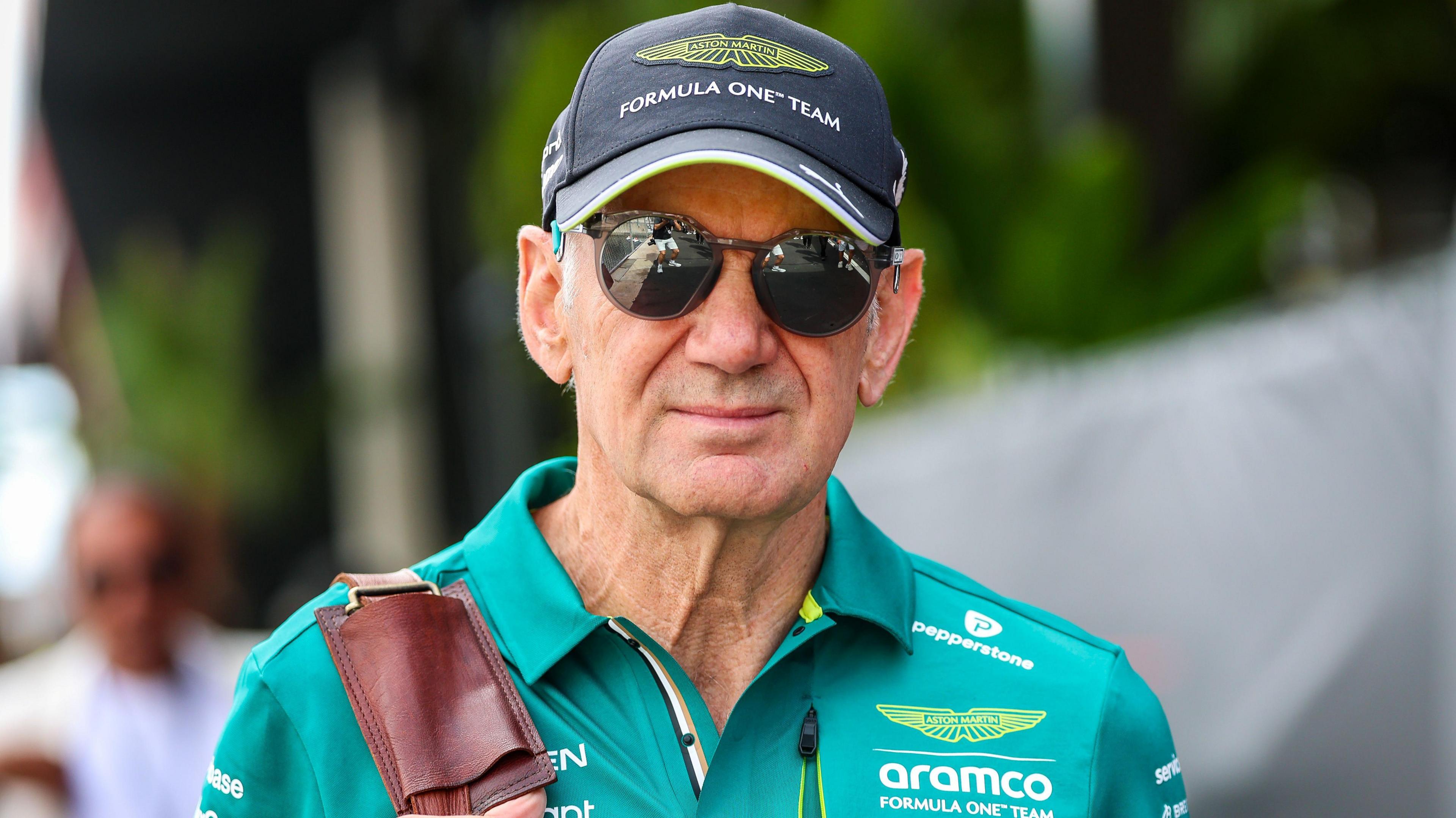 Adrian Newey in the Monaco paddock wearing sunglasses, and an Aston Martin cap and green polo shirt. He is carrying a bag over his right shoulder