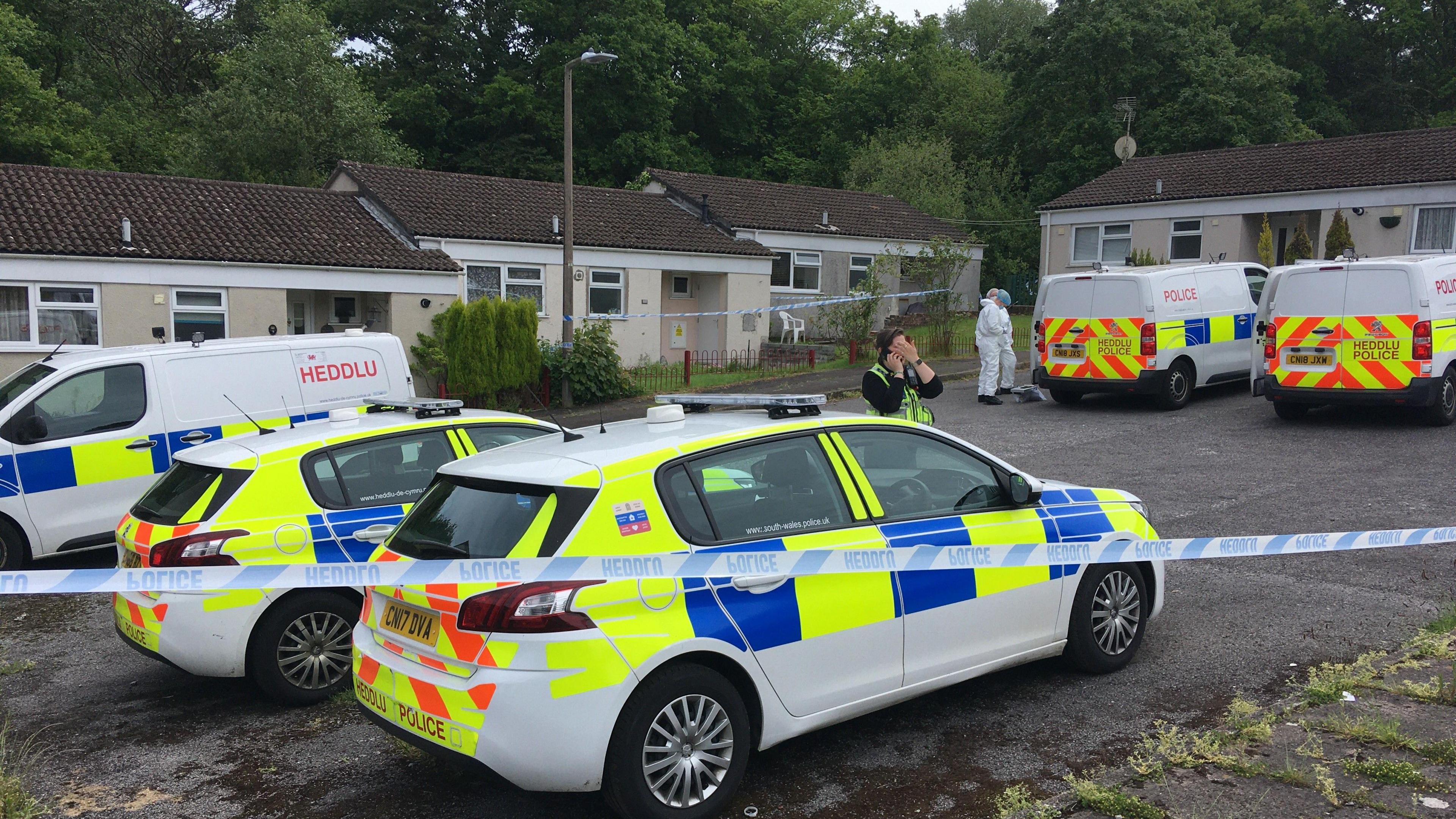 two police cars, three police vans, an officer in uniform and another in a white forensic coverall in front of several bungalow homes including one behind police tap 