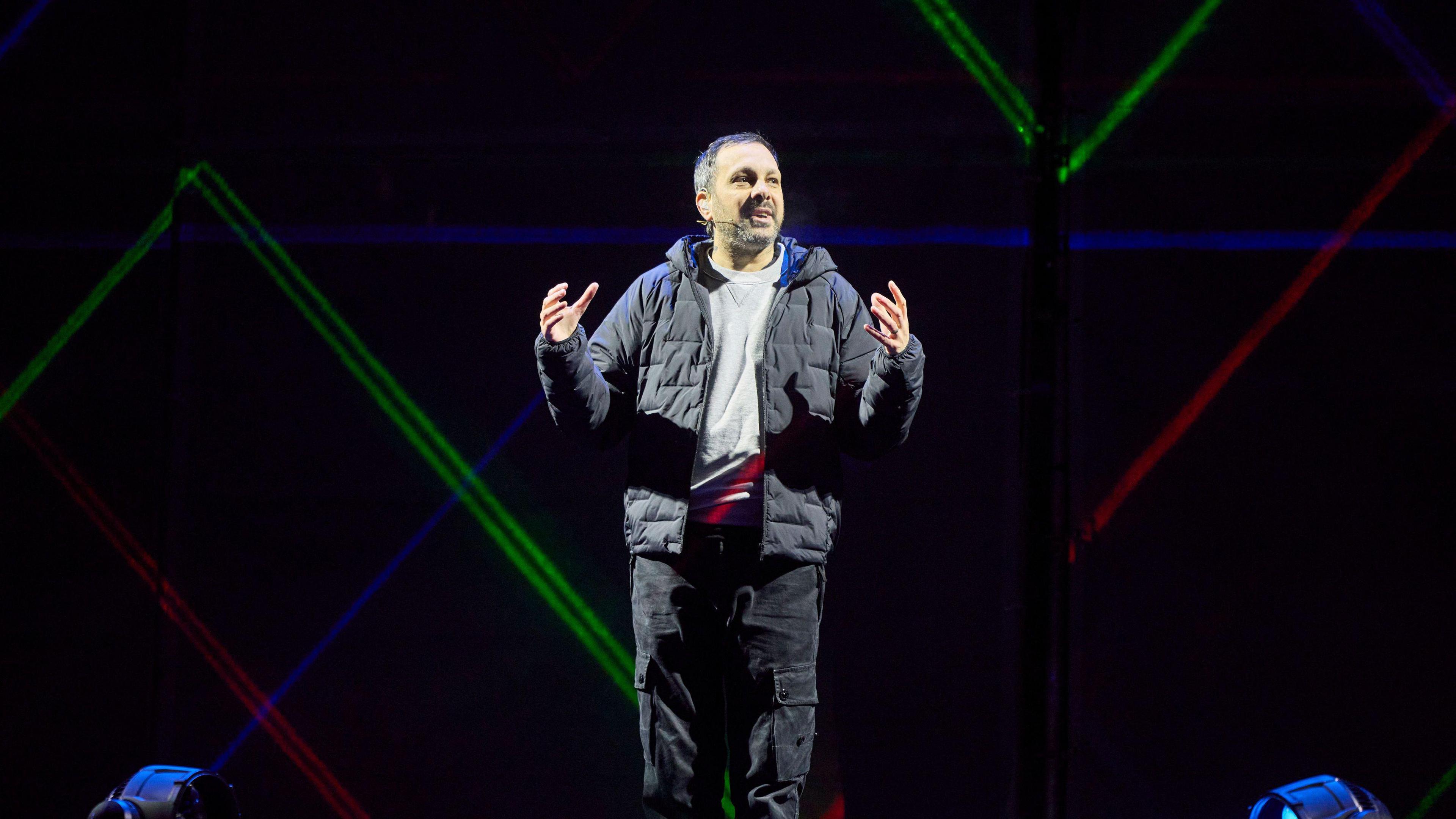 Steven Frayn stands on stage with his hands gesturing he is a man with short brown-grey hair. He is wearing black cargo trousers, a grey jumper and a black puffer jacket. Behind him are coloured lights.