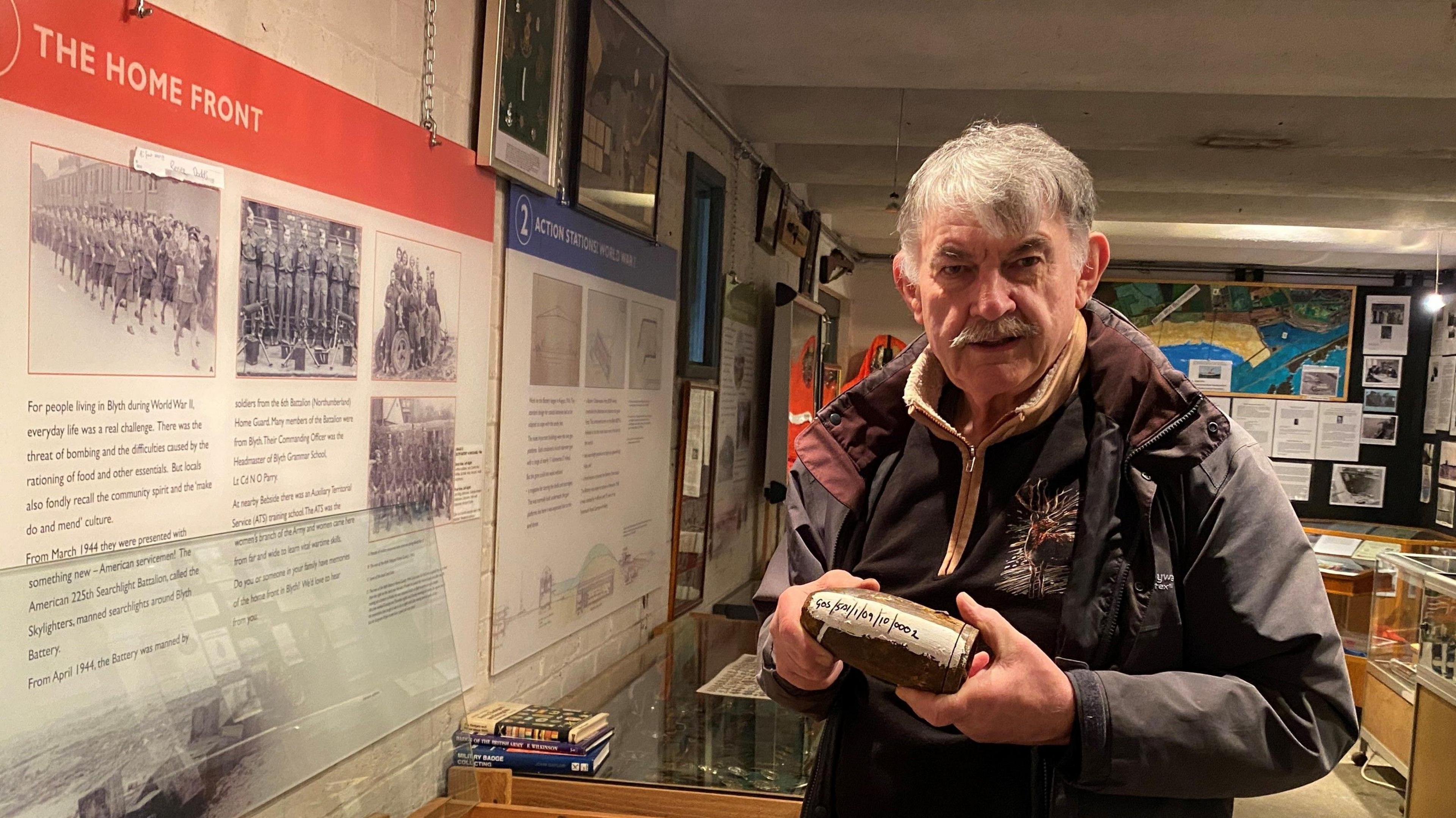 Colin Durward is a man in his 60s who is standing in a museum holding a second world war era bomb. He has grey hair and a moustache and is wearing a brown jacket.