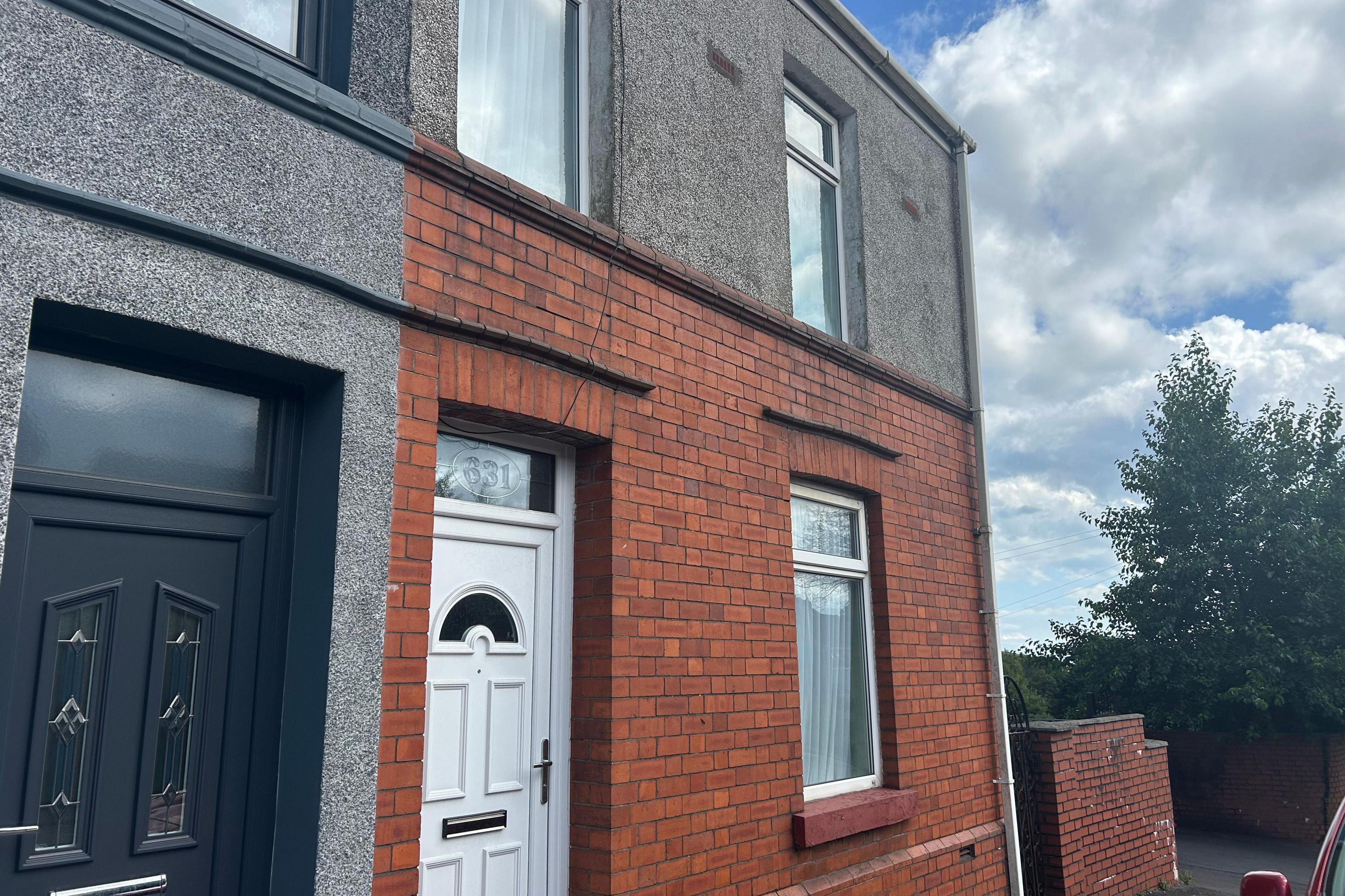 A close up exterior image of the red-brick, two-storey terraced house on the end of the street. Three windows and a white PVC door can be seen. The house next to the red-brick one is pebble-dashed and has a blue PVC door and can be seen on the left of the image. To the right of the house there is a wall and a bush. The sky is blue and there are clouds. 