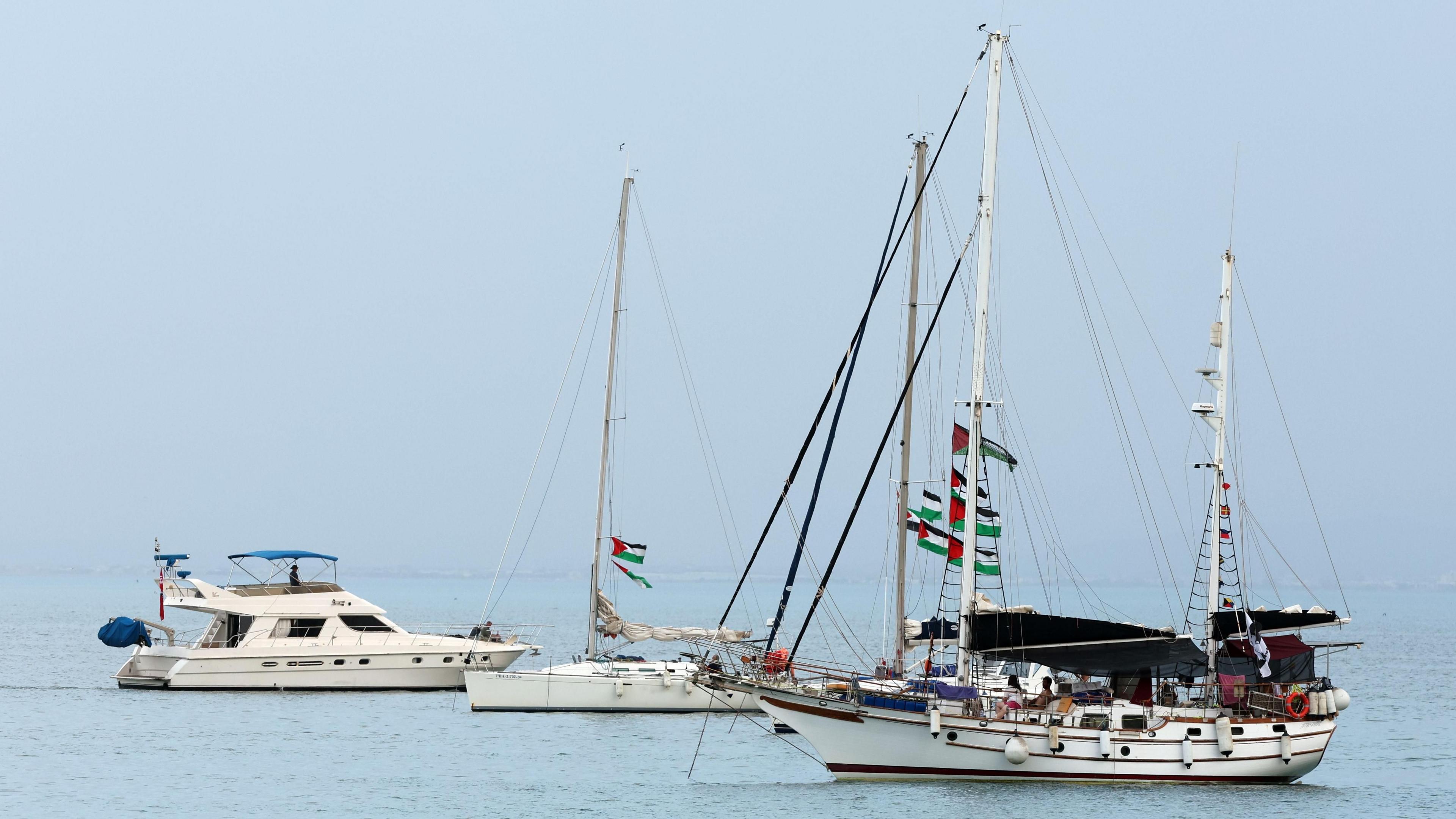 Three sailing boats are seen anchored in turquoise waters, with Palestinian flags strung through the rigging and masts of the vessels.