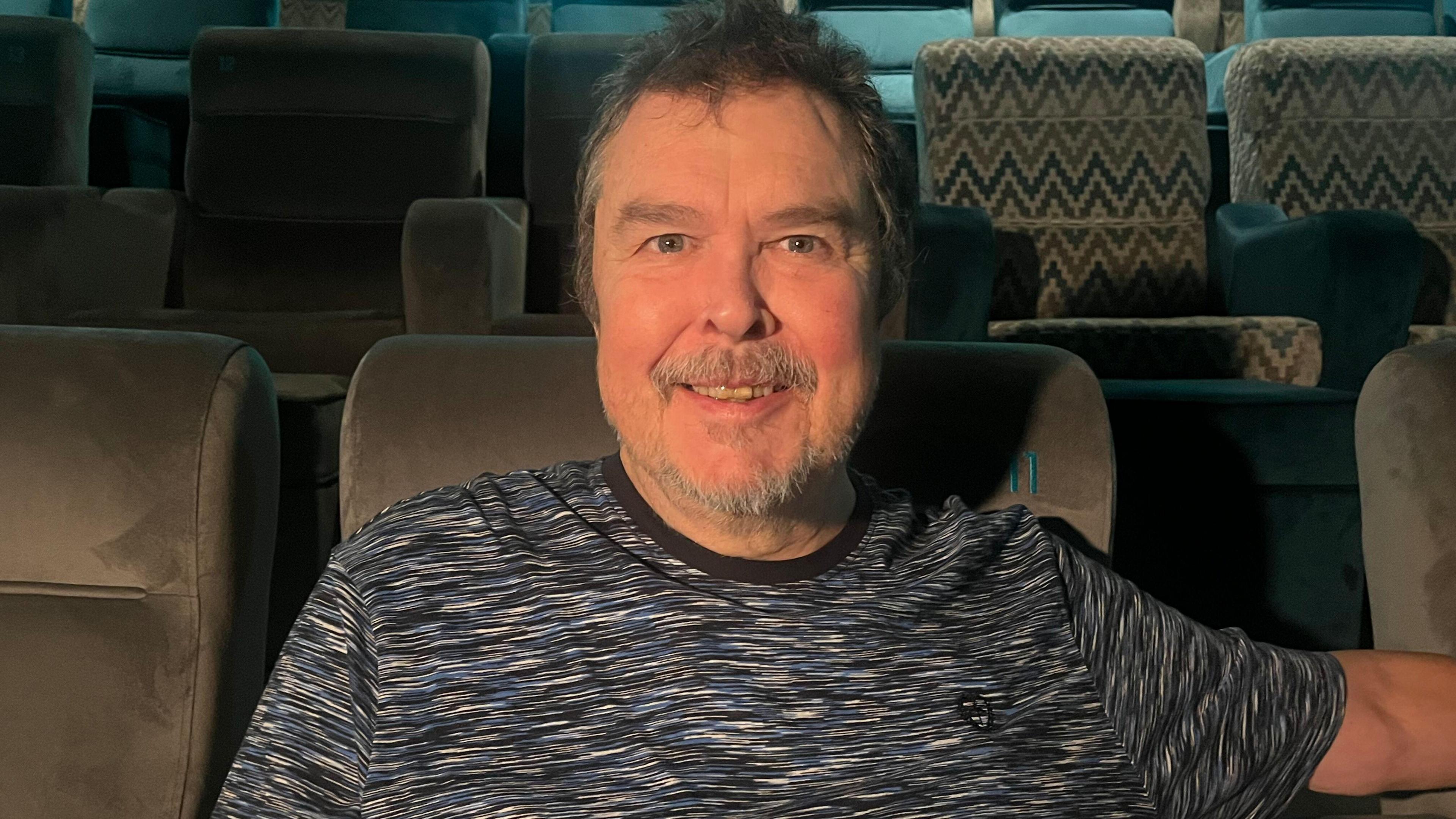 A man with short dark hair, a greying beard and moustache, is sitting in a cinema seat wearing a blue, black and white patterned t-shirt