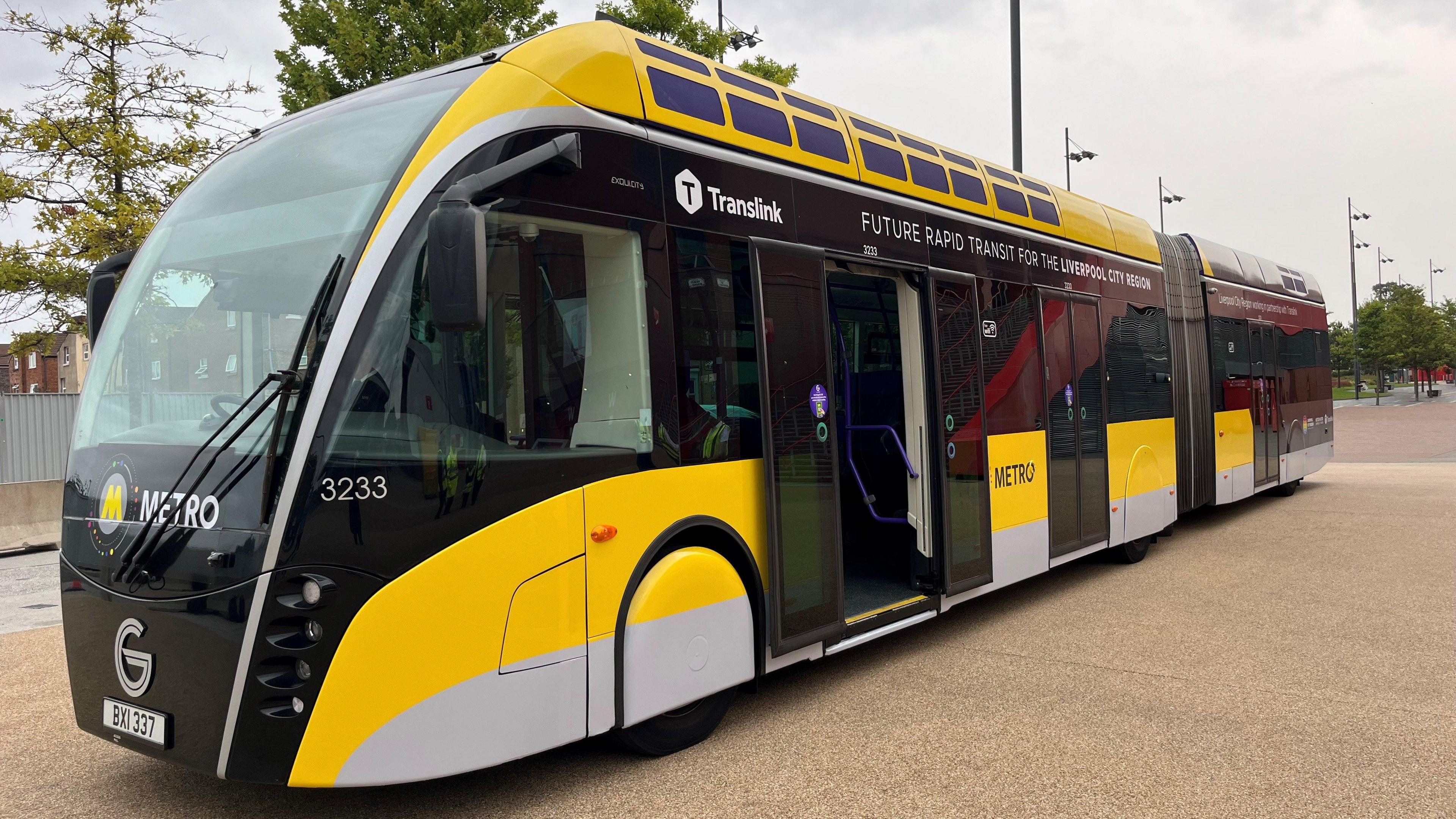 The trial glider bus painted in Merseyrail black and yellow brand colours. It says Future Rapid Transport for Liverpool City Region at the top. 