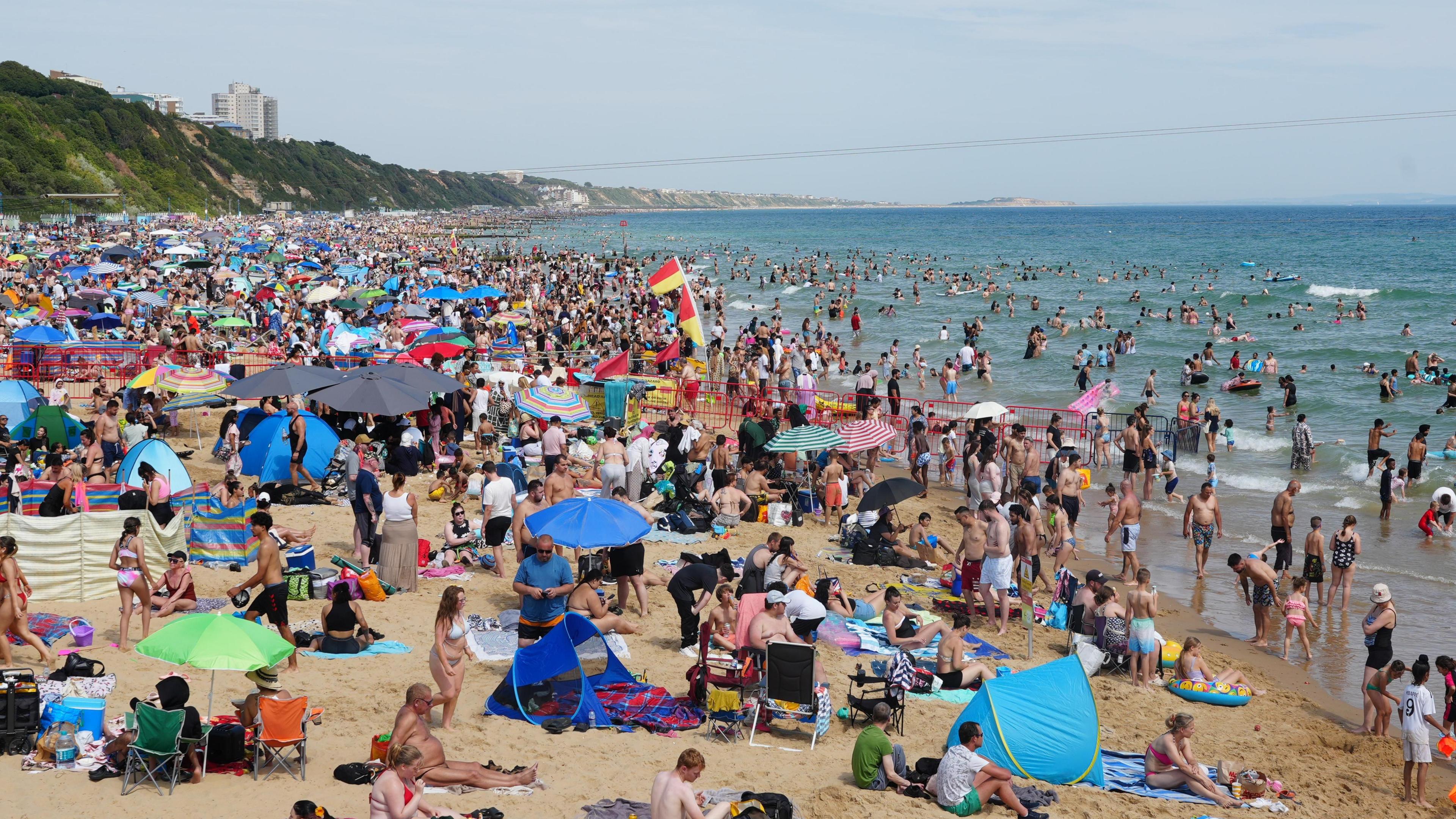 A beach is filled with thousands of sunbathers, parasols and tents as crowds flock to the beach in the sunshine. Many are paddling and swimming in the sea. red and Yellow lifeguards flags can be seen standing in the sand.