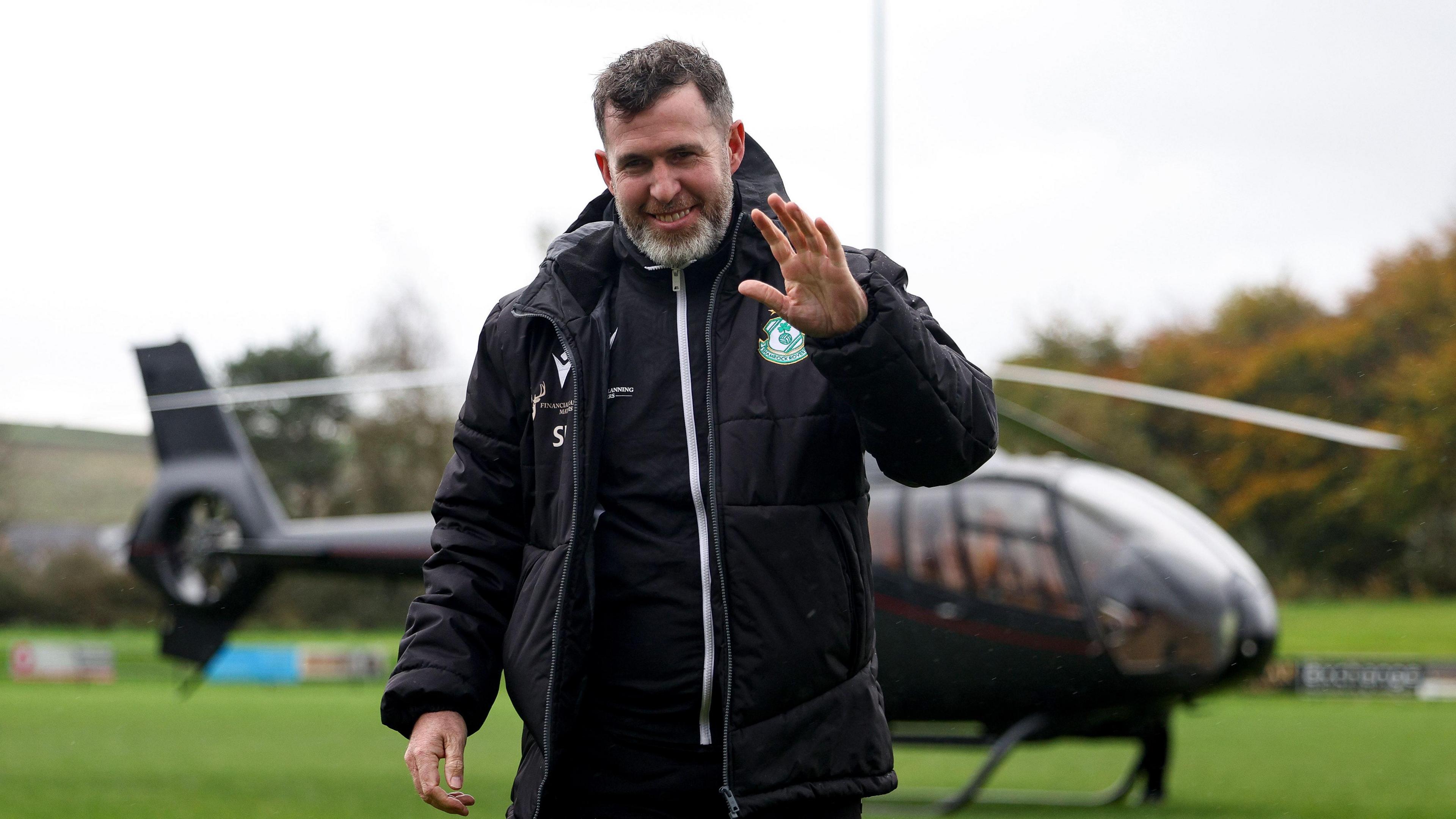 A man in a black sports coat with a green and white football crest waves to the camera. There is a black helicopter behind him on a grass field. There are also trees in the background.