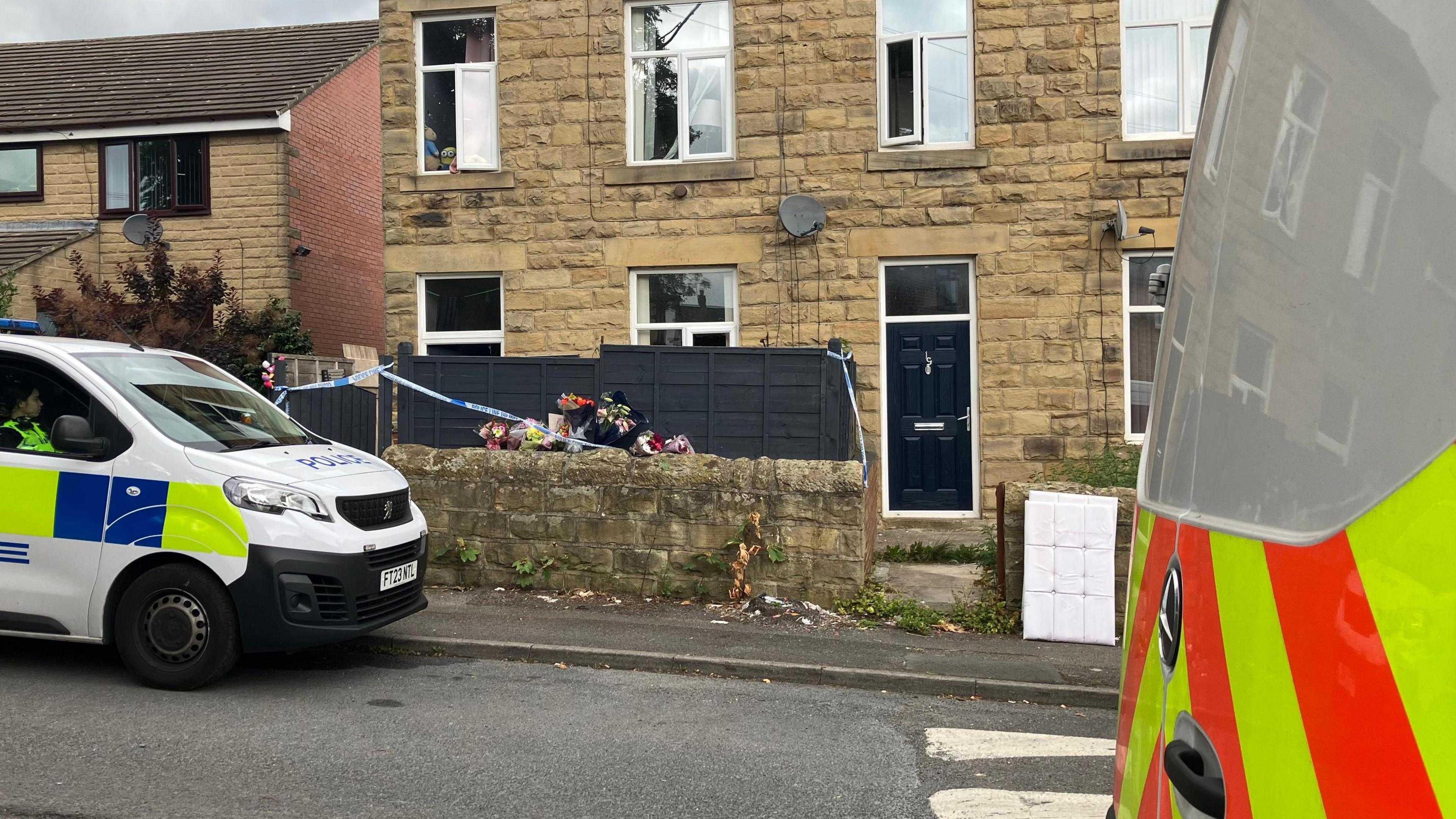 Two police vans parked either side of a terraced house. Police cordon tape around the front of the house and bunches of flowers on the wall outside the house.