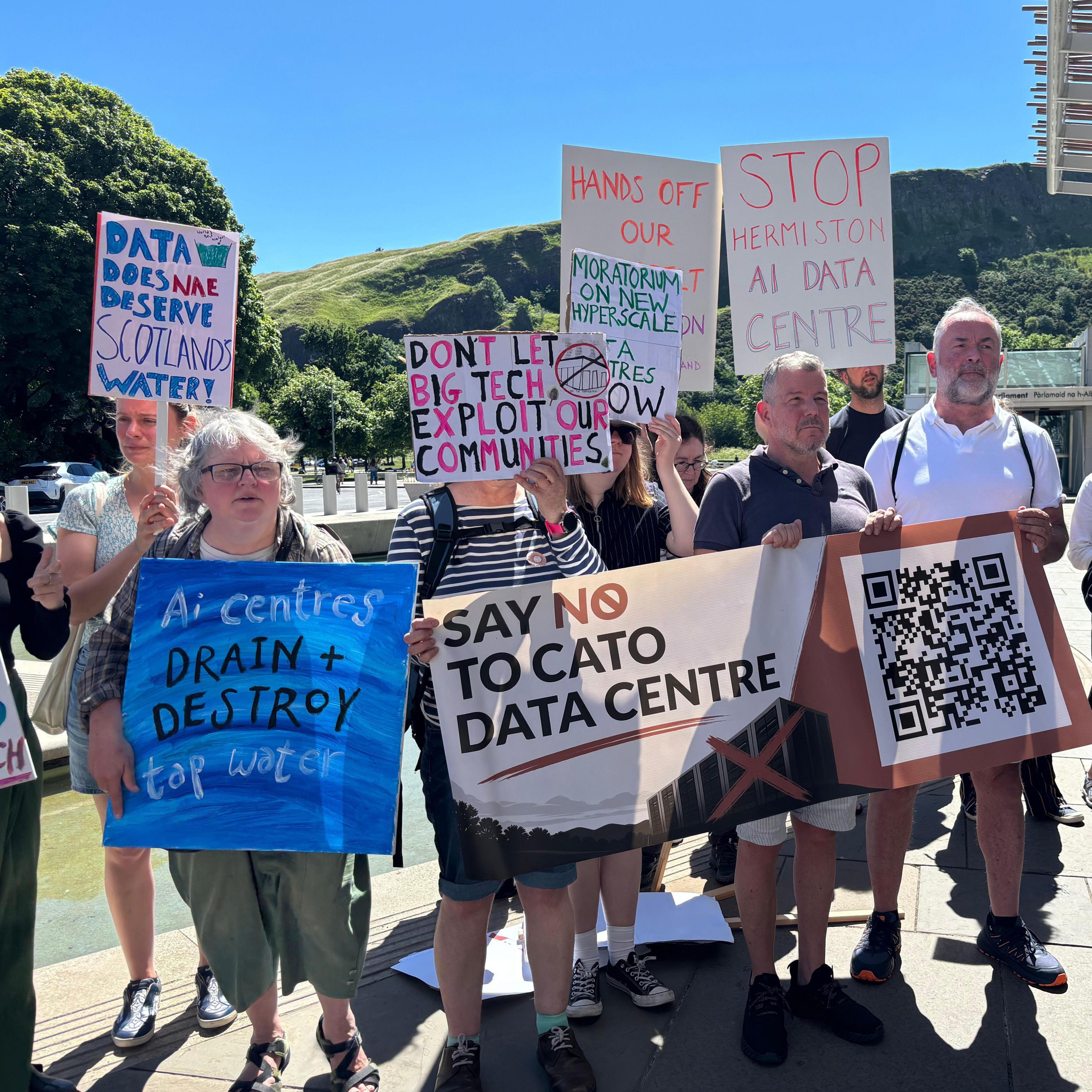 A group of people stand outdoors in bright sunshine holding protest signs opposing a data centre development. The group is gathered near a modern building with a metal canopy, with green hills and a clear blue sky in the background.