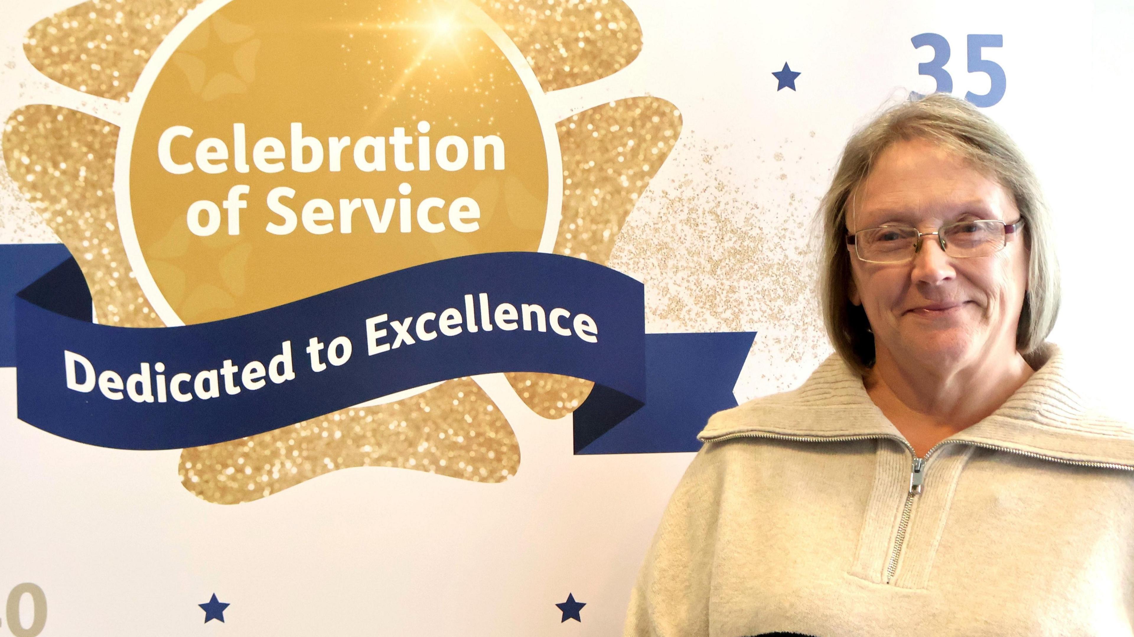 Mandy Lovell is smiling at the camera in front of a backdrop that says "celebration of service" and "dedicated to excellence". She has fair bob-length hair and is wearing spectacles and a beige pullover with a zip at the front