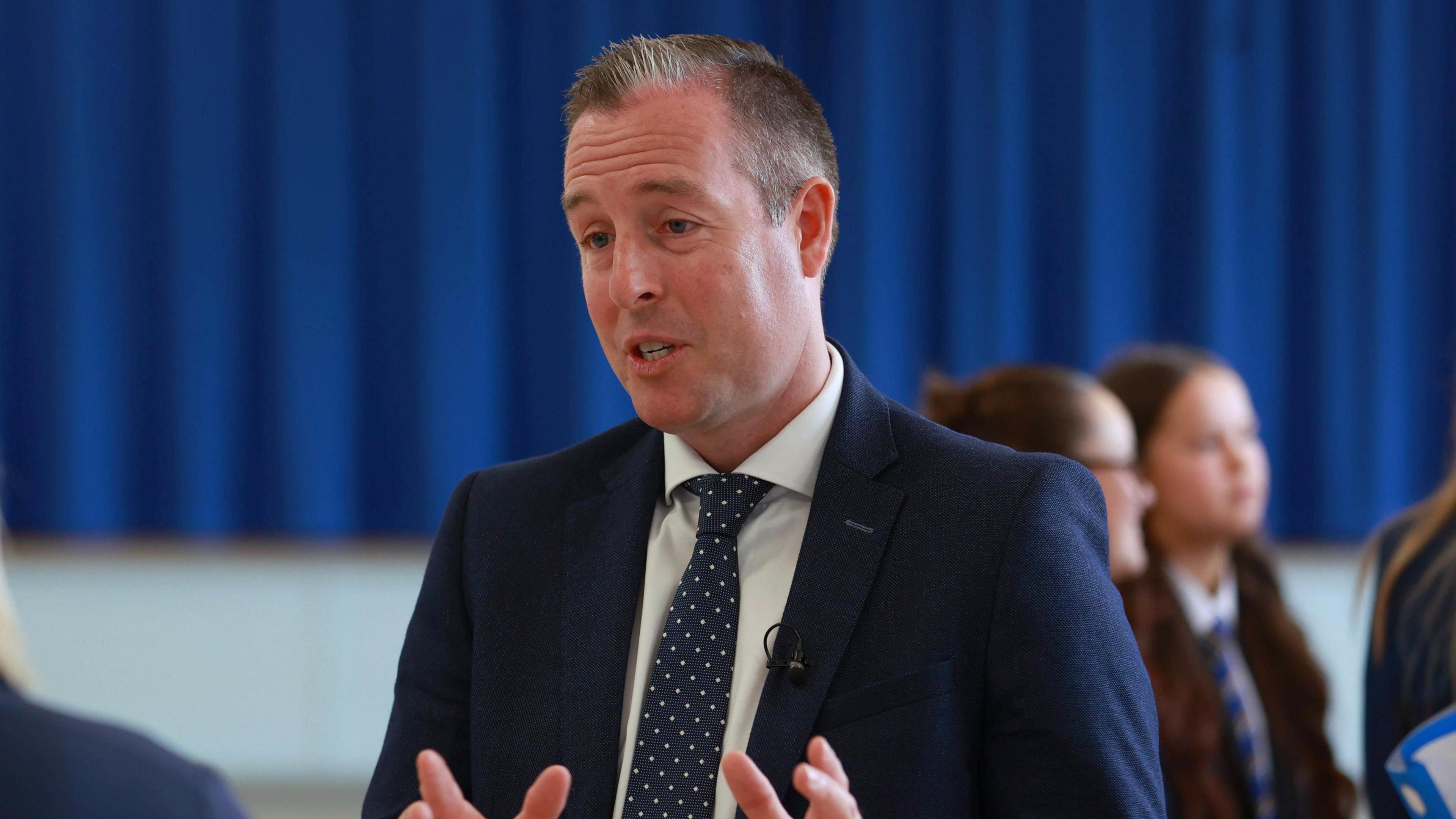 A man wearing a navy suit and tie is pictured with his hands out as he seems to be in mid-conversation. Some pupils are gathered in the background in front of a blue curtain. 