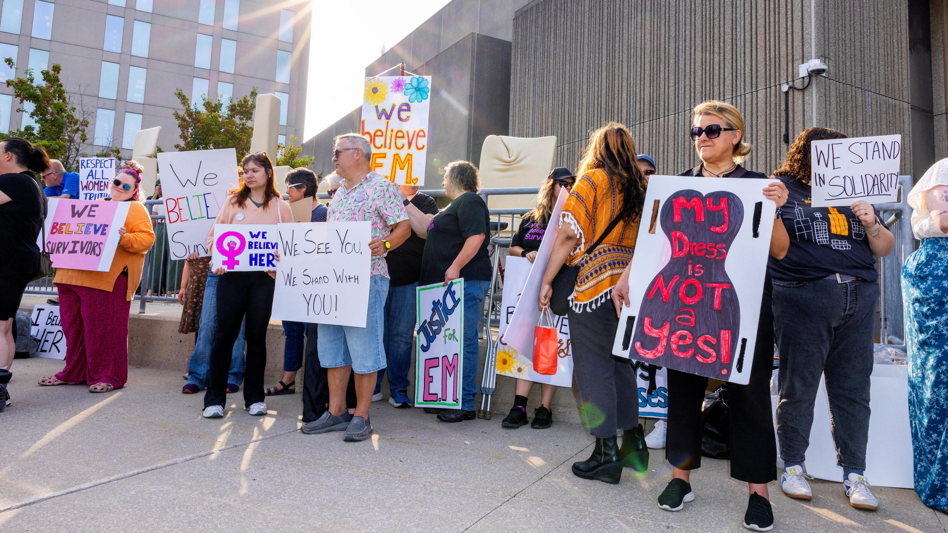 Protesters in front of courthouse hold signs showing support for EM that read "We believe her", "Justice for EM" and "My dress is not a yes"