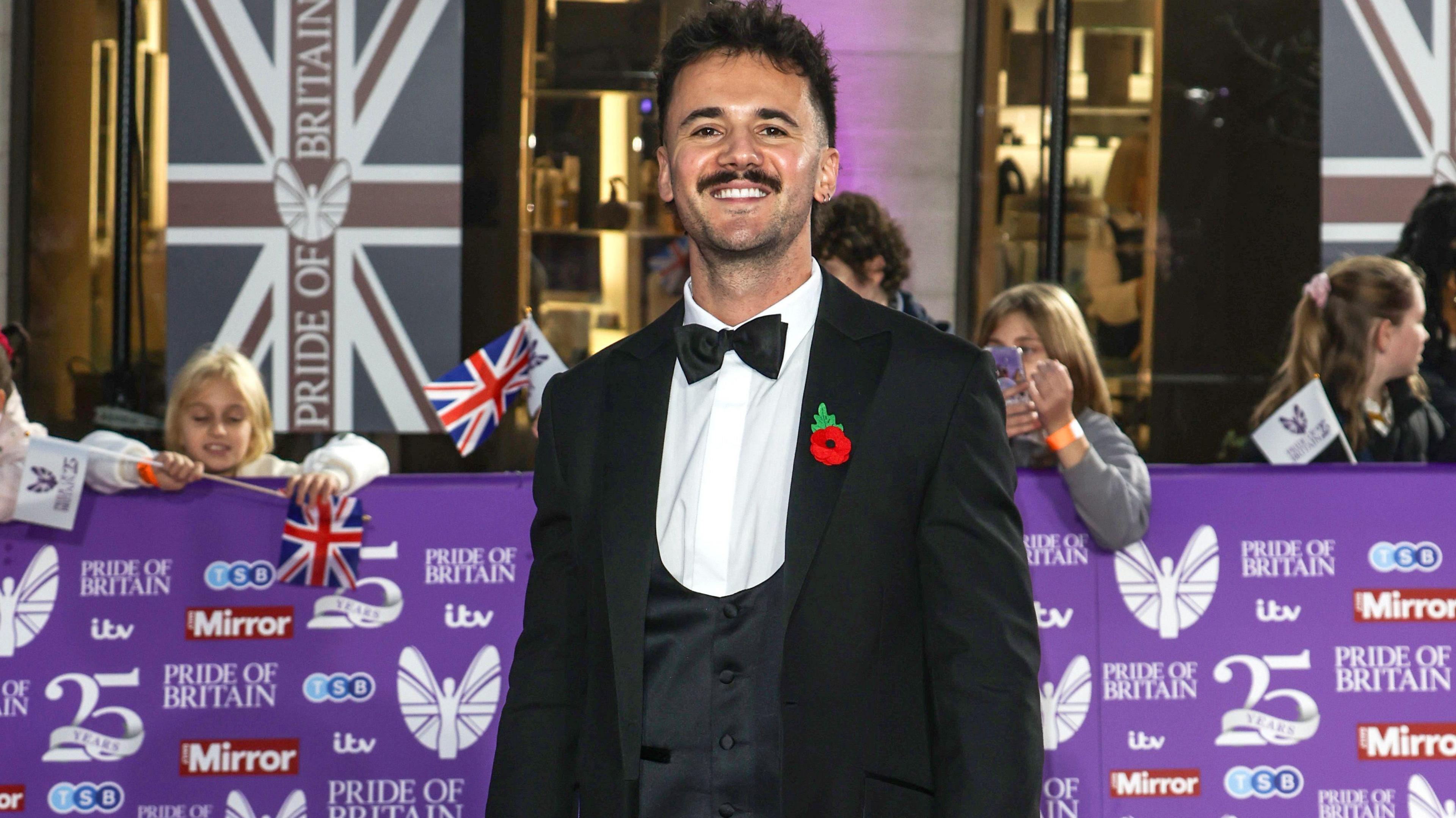 Luke at the Pride of Britain Awards wearing a blue suit, with white shirt and black bowtie, he has a red poppy on his lapel and there are people behind him. He is smiling, with a moustache and is looking straight at the camera. There is a purple banner behind him and Pride of Britain banner, dropping down.