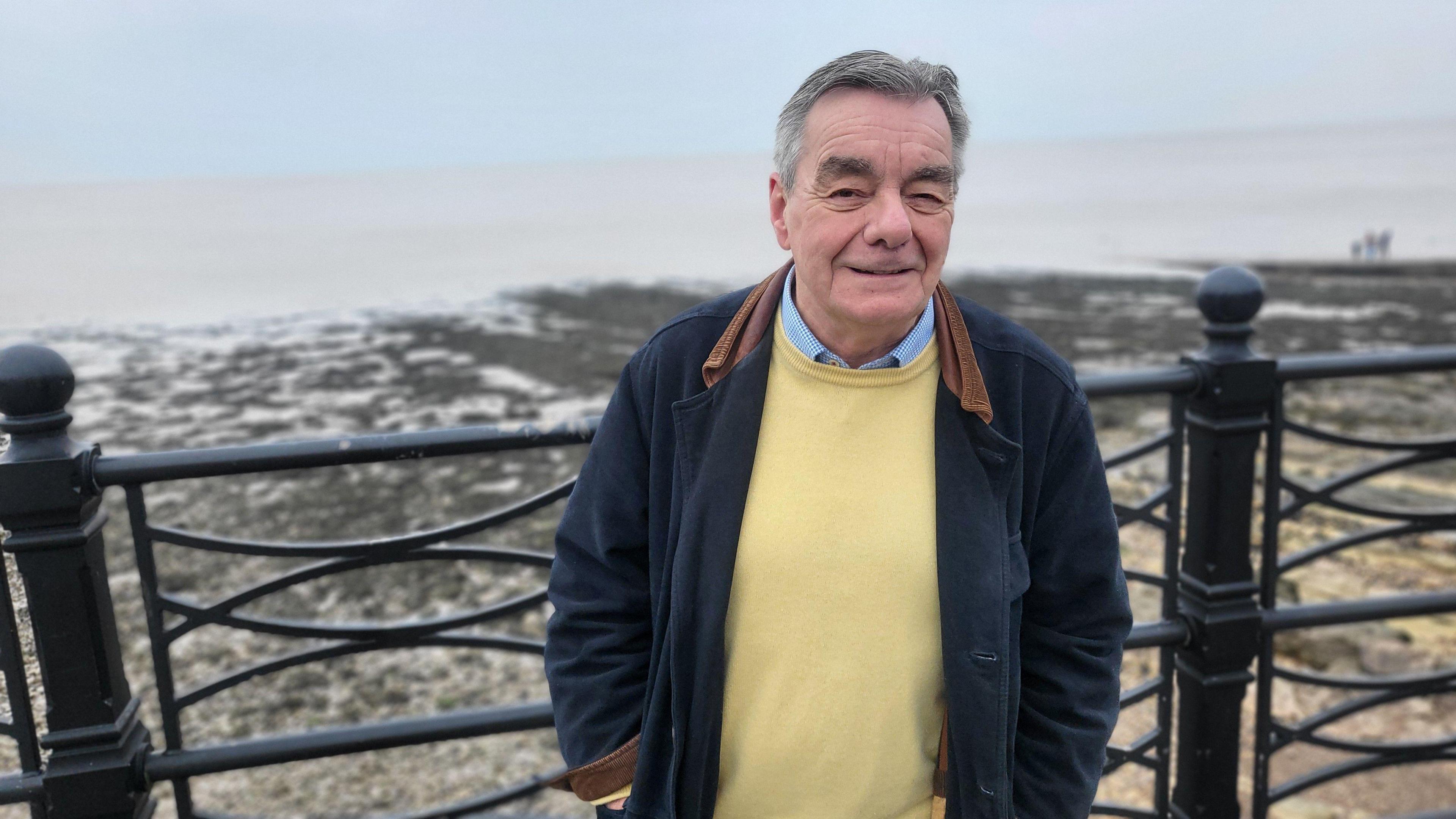 A man wearing a navy jacket and yellow jumper is standing for a photo in front of Clevedon seafront. The sea is behind him.