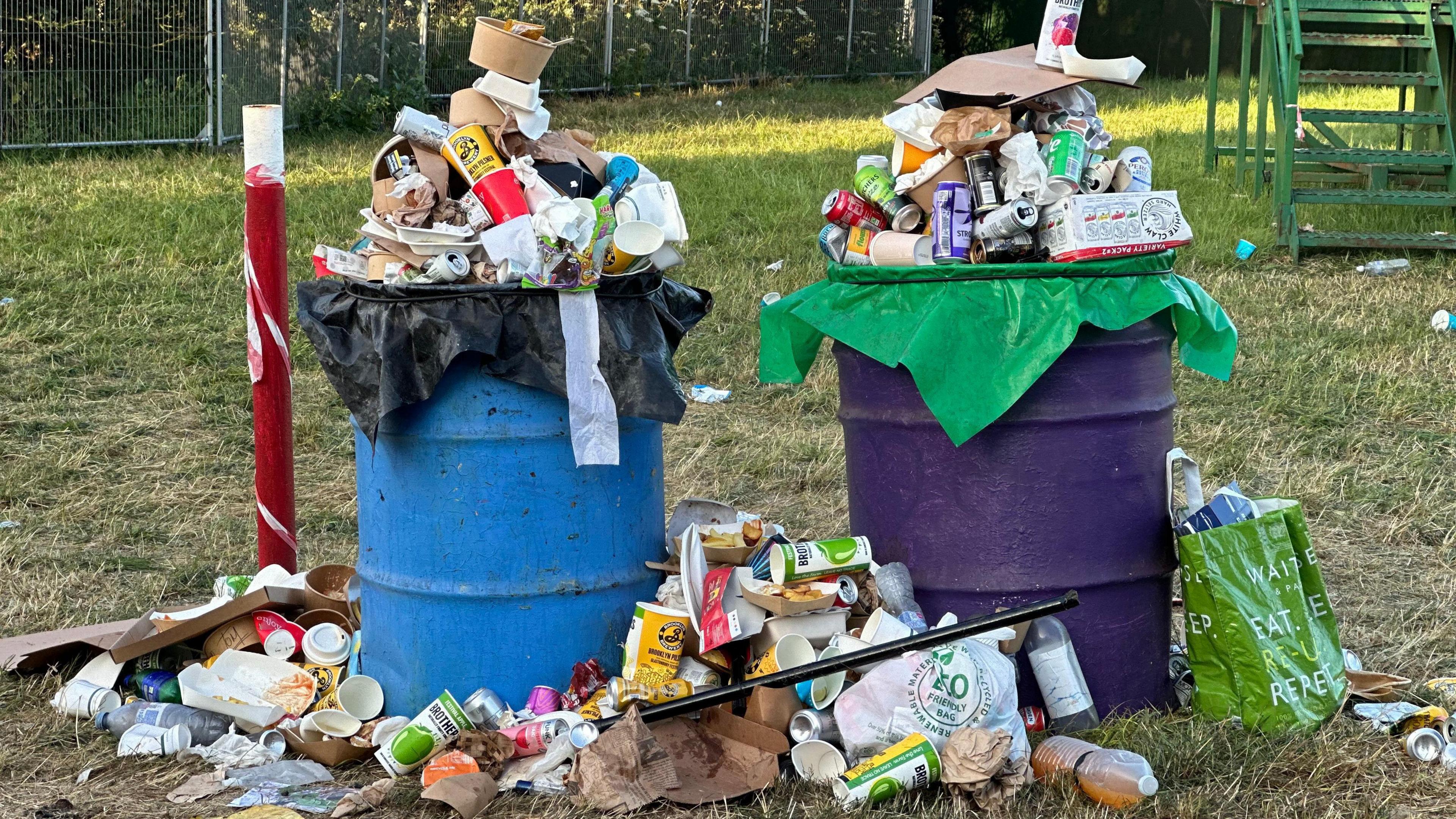 Two overflowing bins at Glastonbury Festival. There is rubbish on the floor surrounding them too.