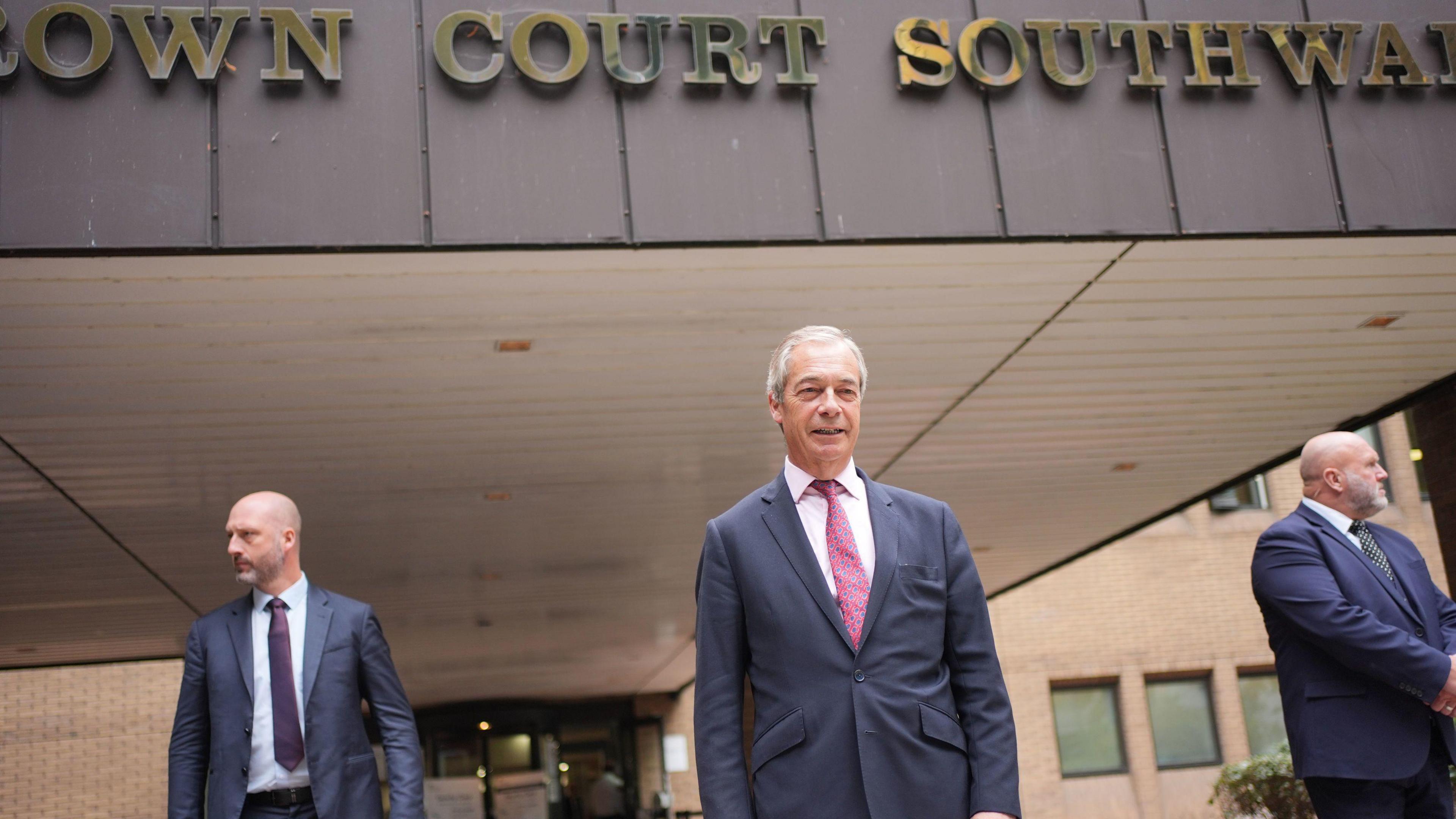 Nigel Farage wearing a suit standing in front of a building that says Crown Court Southwark. There are two other men in the picture standing slightly behind him.