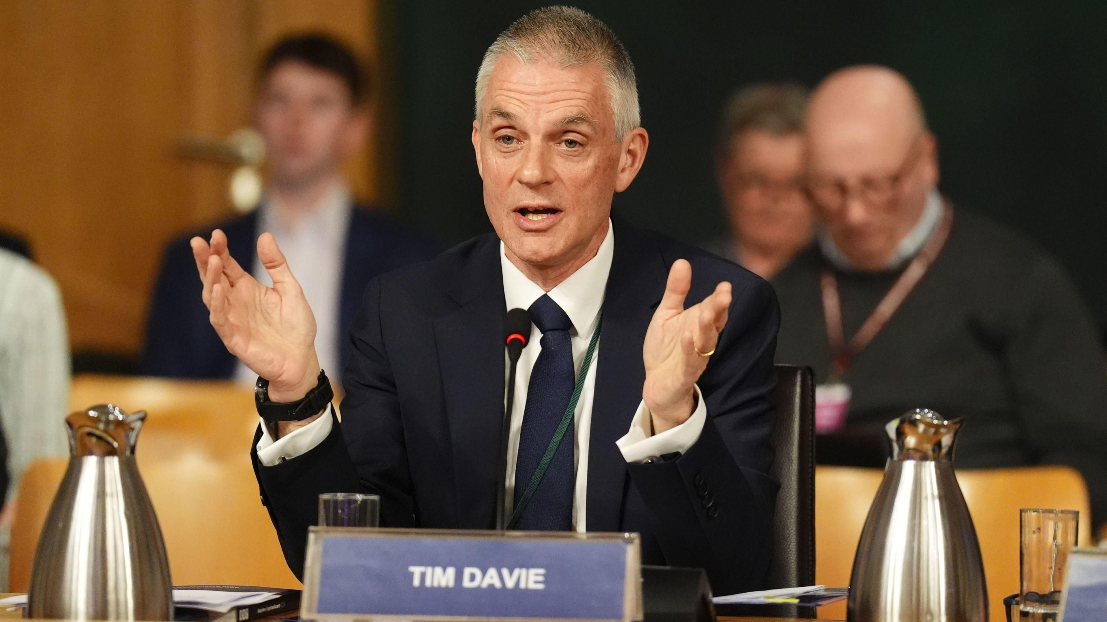 Tim Davie, sitting on a panel, wearing a suit and gesturing with his hands as he talks