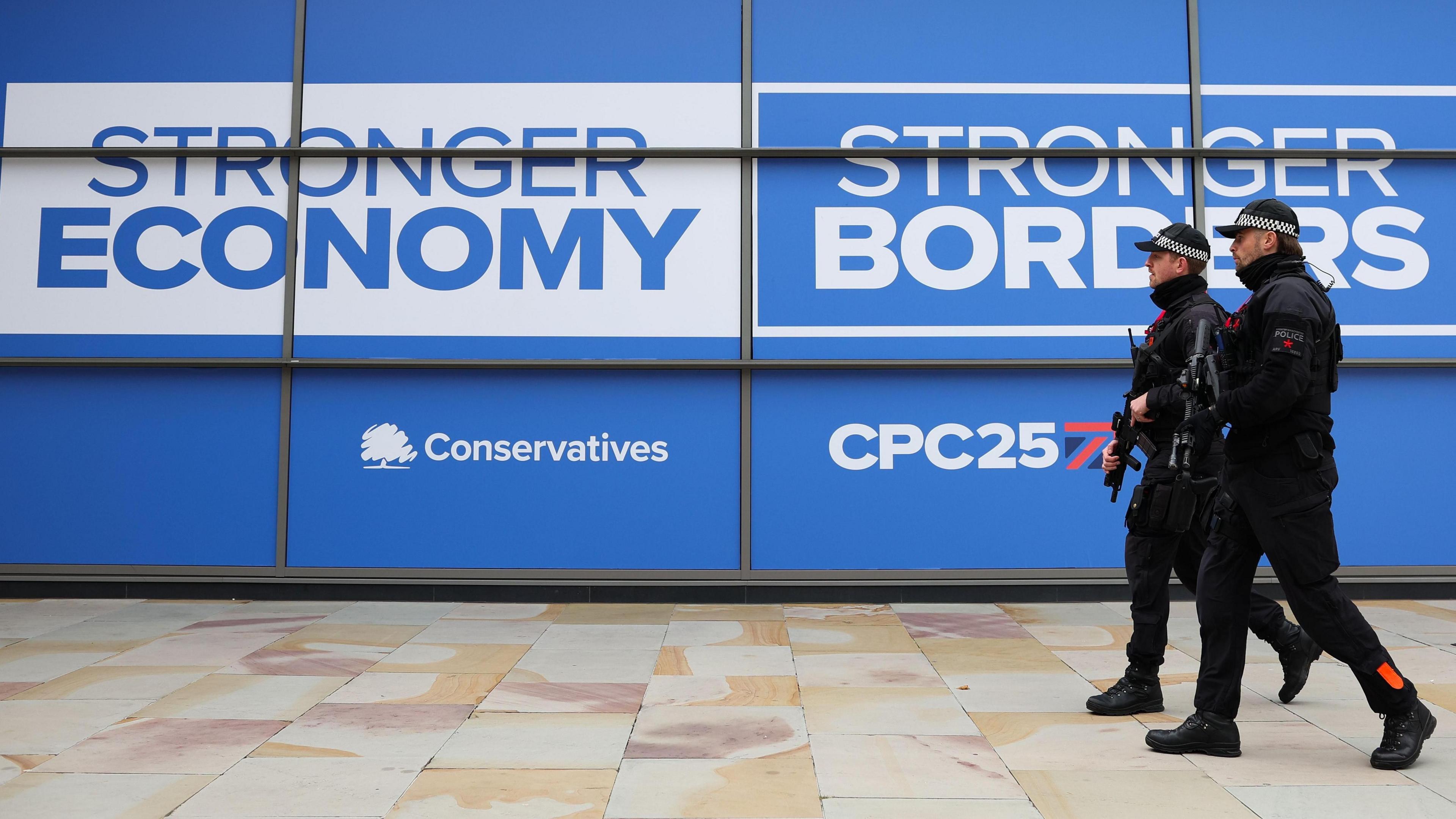 Two police officers with guns walk in front of a blue Conservatives sign reading 'Stronger economy stronger borders'
