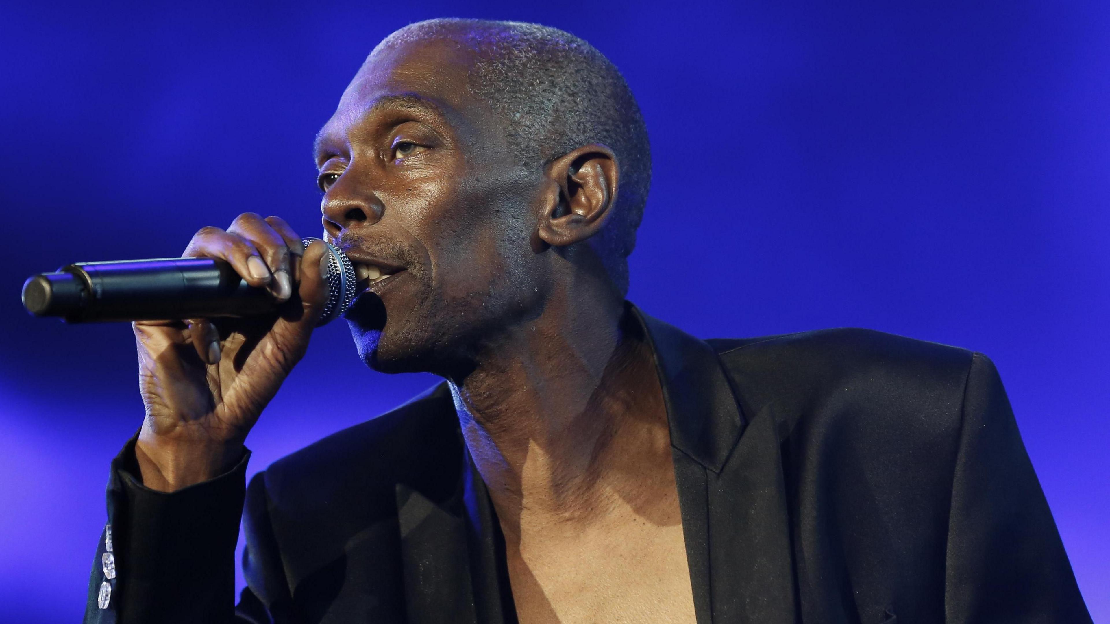 A man is pictured singing into a microphone he is holding in his right hand. He is wearing a black blazer with no shirt underneath.