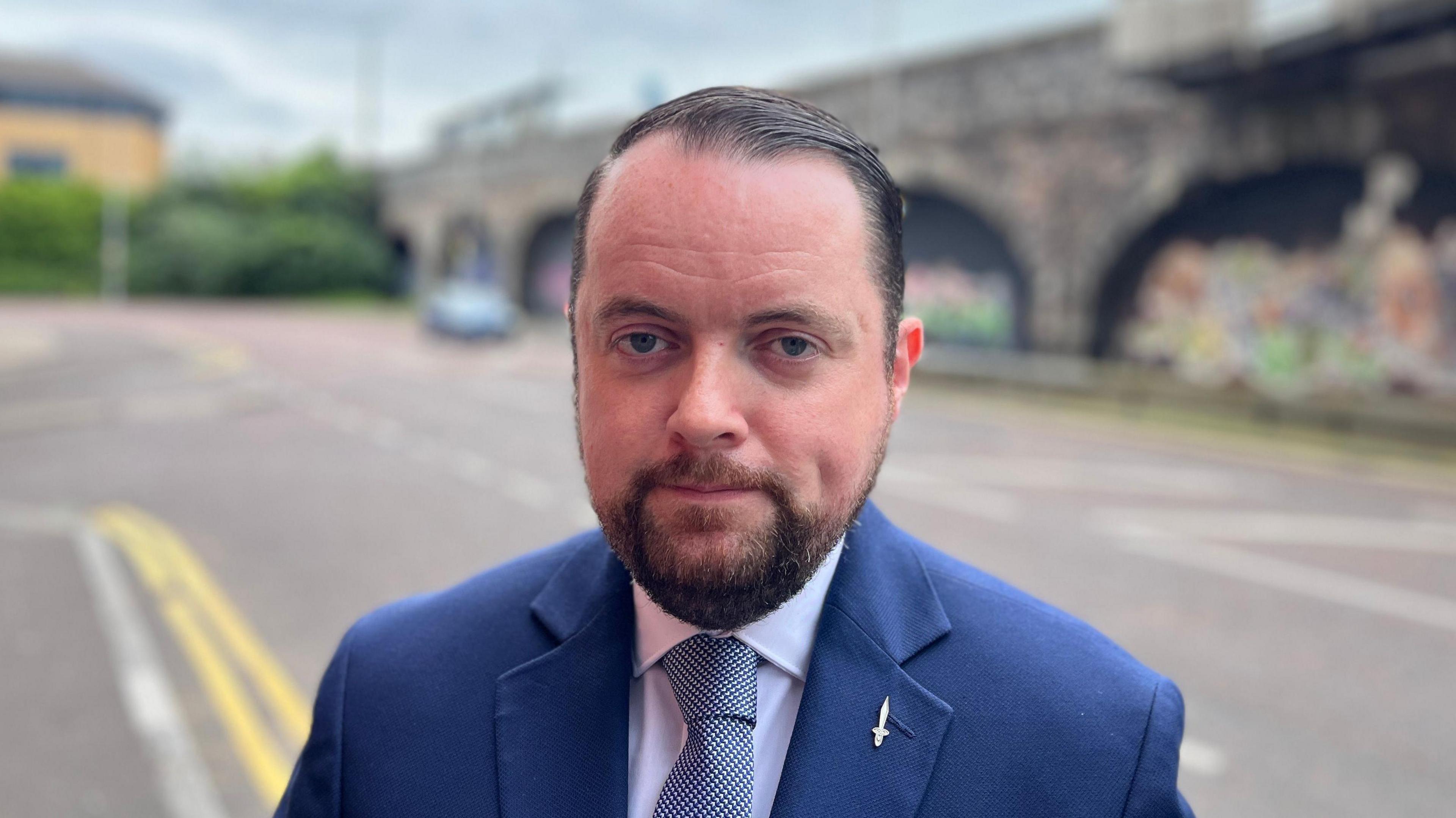 Fionntán Hargey is wearing a white shirt, navy blazer and a white and blue tie. He has black hair and a black beard. On his blazer is a silver pin of a sword. The background is blurred but we can see a bridge, a car and double yellow lines alongside the footpath.