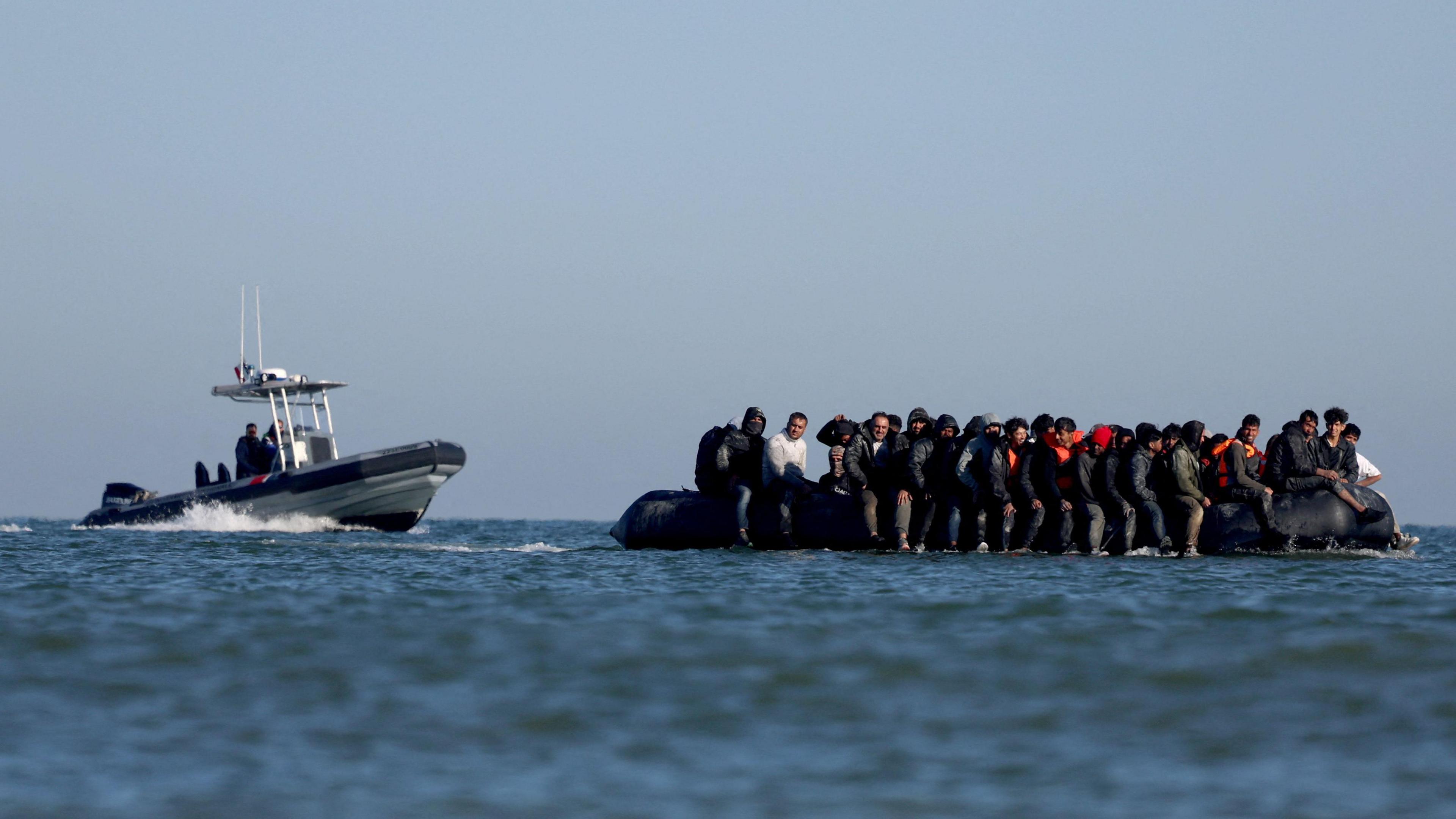 A group of migrants, some of them wearing safety vests, are sitting on an inflatable dinghy at sea. A French police boat is approaching them from behind. The sky is blue and the sea is relatively calm.
