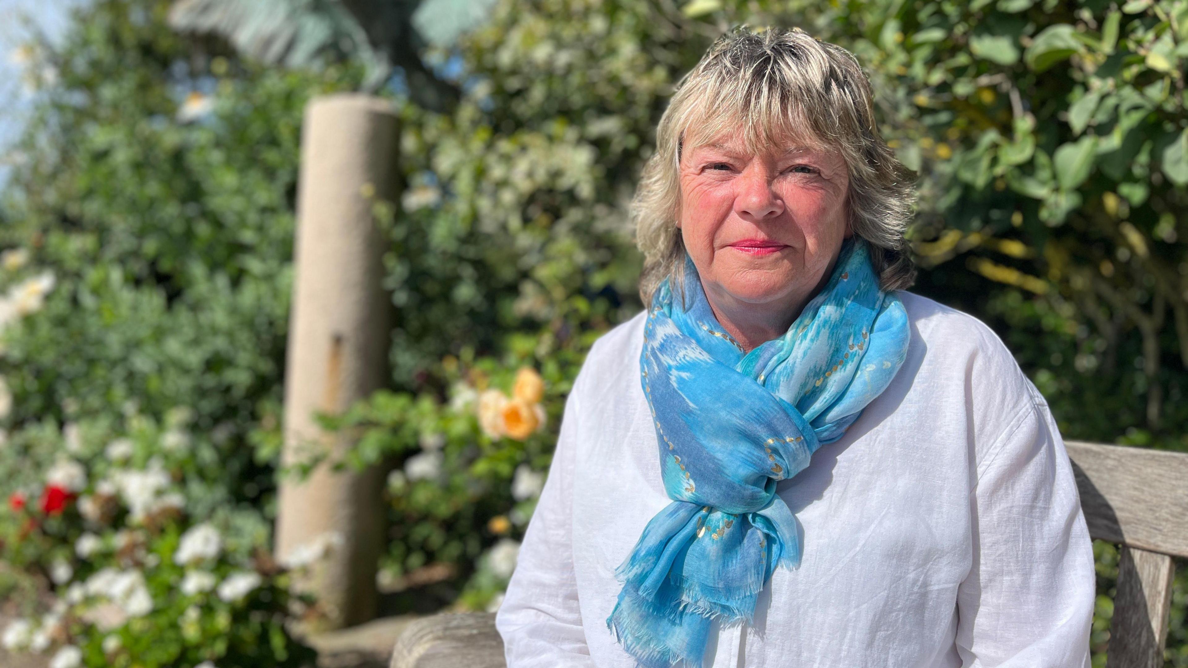 Alison looks at the camera as she sits on a bench and there are flowers in a garden in the background. She wears a white blouse and a light blue silk scarf.